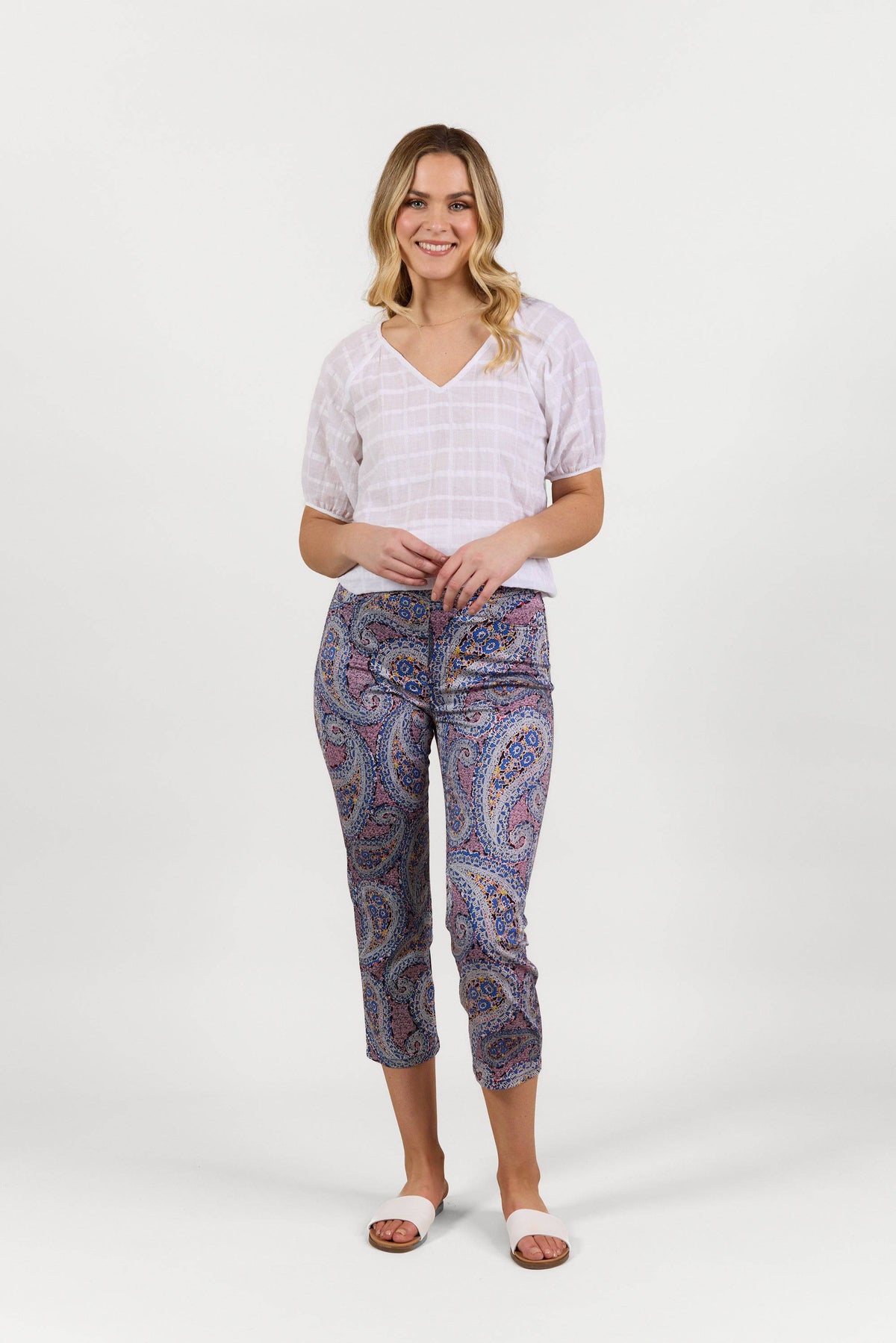 A woman stands smiling against a plain white background, wearing a white short-sleeve V-neck top, Vassalli Sierra 7/8 Pant - Paisley, and white slide sandals.