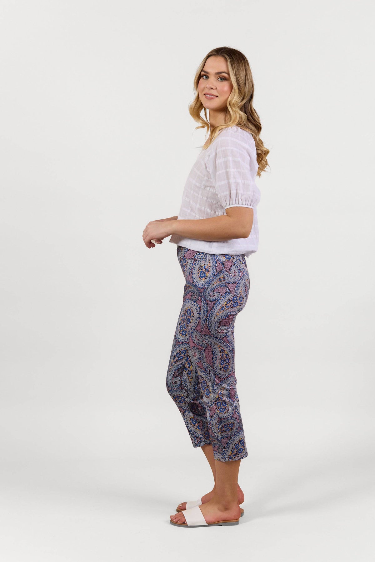 A woman with long wavy blonde hair stands sideways, smiling slightly, wearing a white short-sleeve blouse, Vassalli Sierra 7/8 Pant - Paisley, and white slide sandals. The colorful pants feature a paisley print and have a lightweight pull-on style.