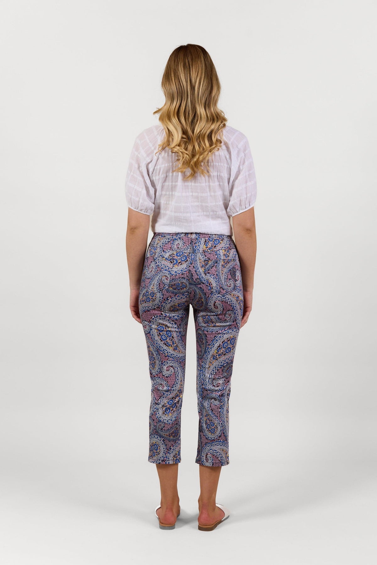 A woman with long wavy blonde hair, wearing a white short-sleeve blouse and Vassalli Sierra 7/8 Pant - Paisley, stands barefoot facing away on a plain white background.