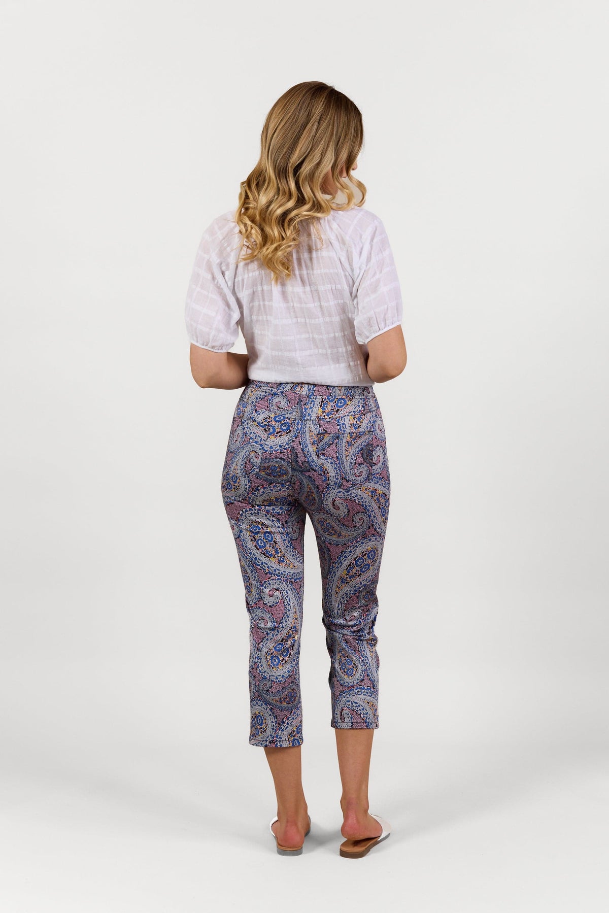 A woman with wavy blonde hair faces away, wearing a white short-sleeve blouse, Vassalli Sierra 7/8 Pant - Paisley, and beige sandals against a plain white background.