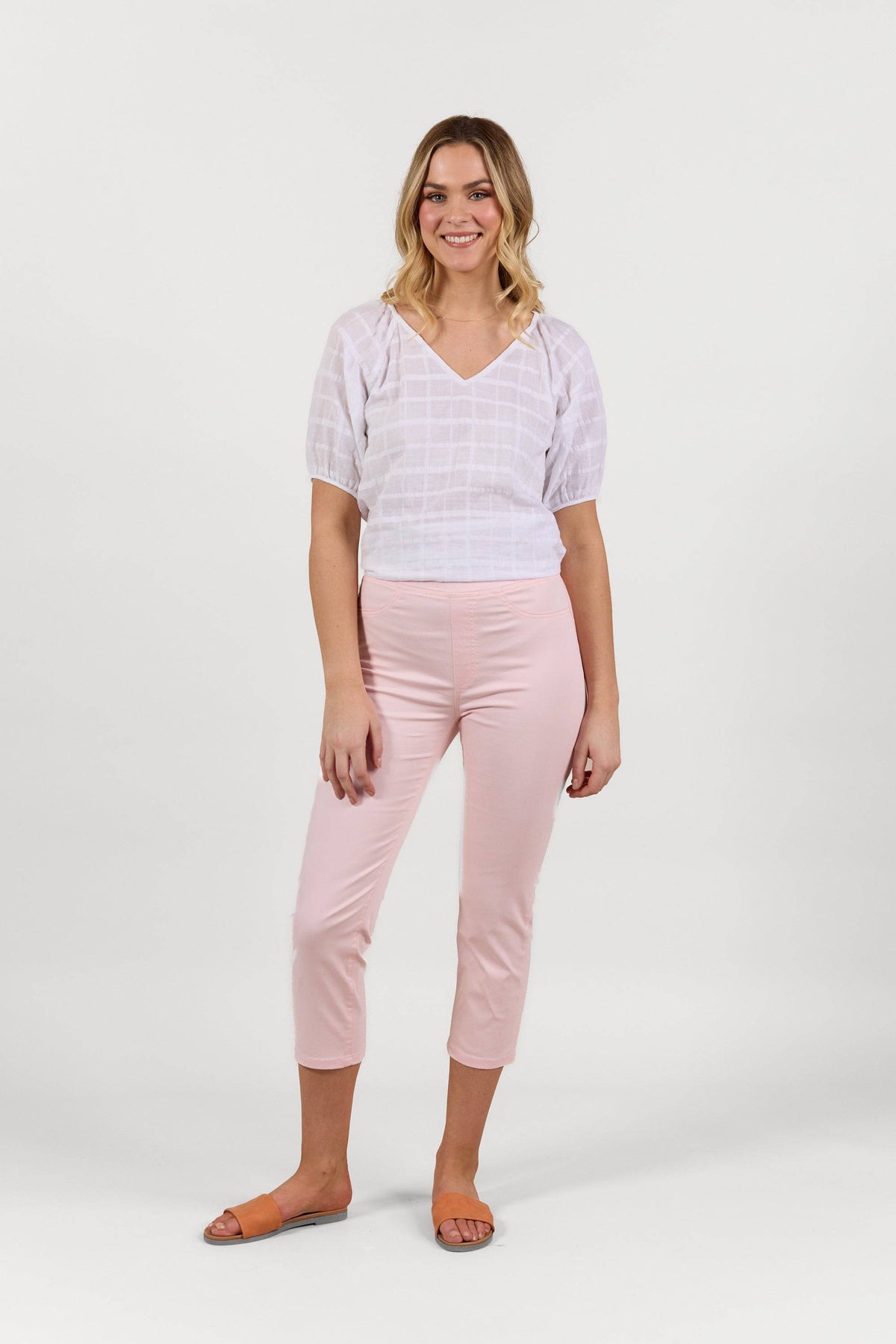 A woman smiles against a white background, wearing a white short-sleeve blouse, Vassalli Sierra 7/8 Pant in Powder Pink, and brown slide sandals.