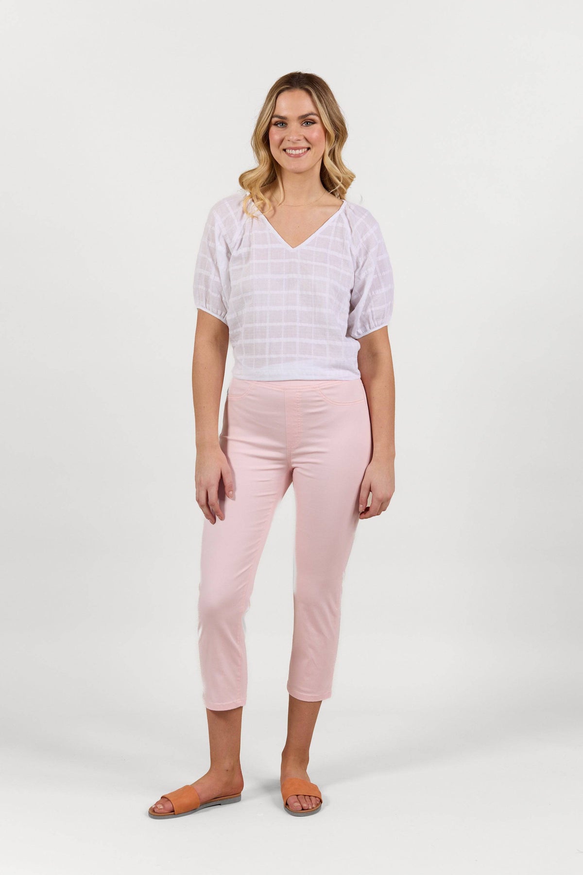 A woman with loose waves smiles against a white background, wearing Vassalli's Sierra 7/8 Pant in Powder Pink, a white short-sleeve top, and tan sandals in a relaxed pose.