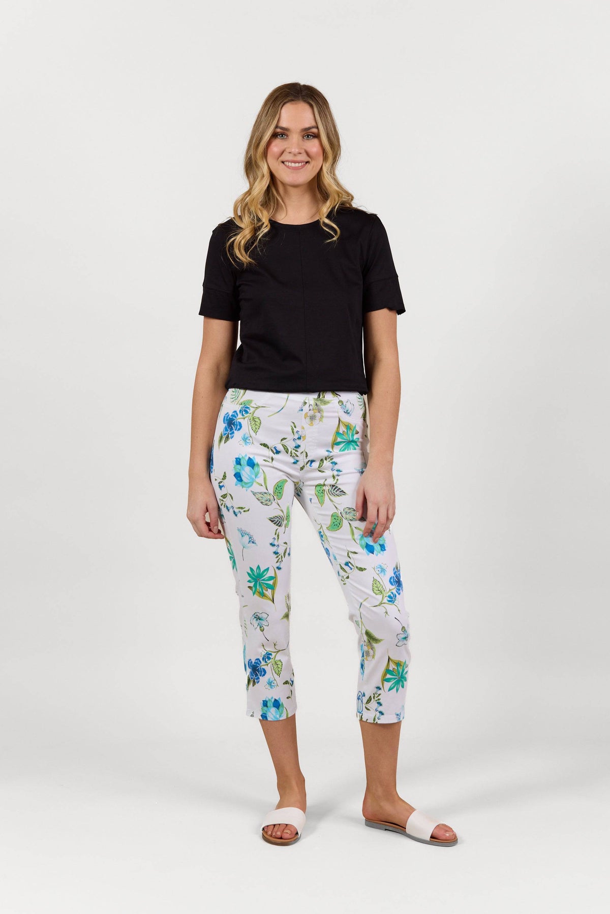 A woman with wavy blonde hair smiles against a white background, wearing a black short-sleeve top, Vassalli Sierra 7/8 Pant - Santorini with a blue and green floral print in a slim leg cut, and white open-toe sandals.