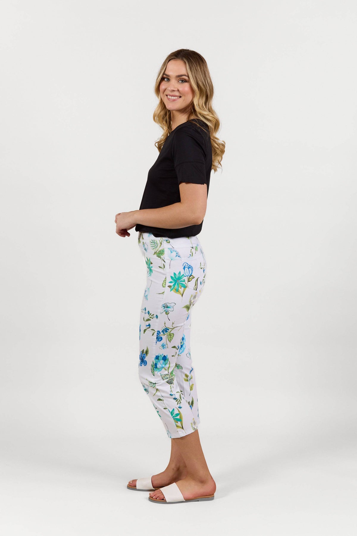 A woman stands sideways, smiling, wearing a black t-shirt and Vassalli Sierra 7/8 Pant - Santorini with a blue and green floral slim leg design, paired with white slide sandals, against a plain white background.