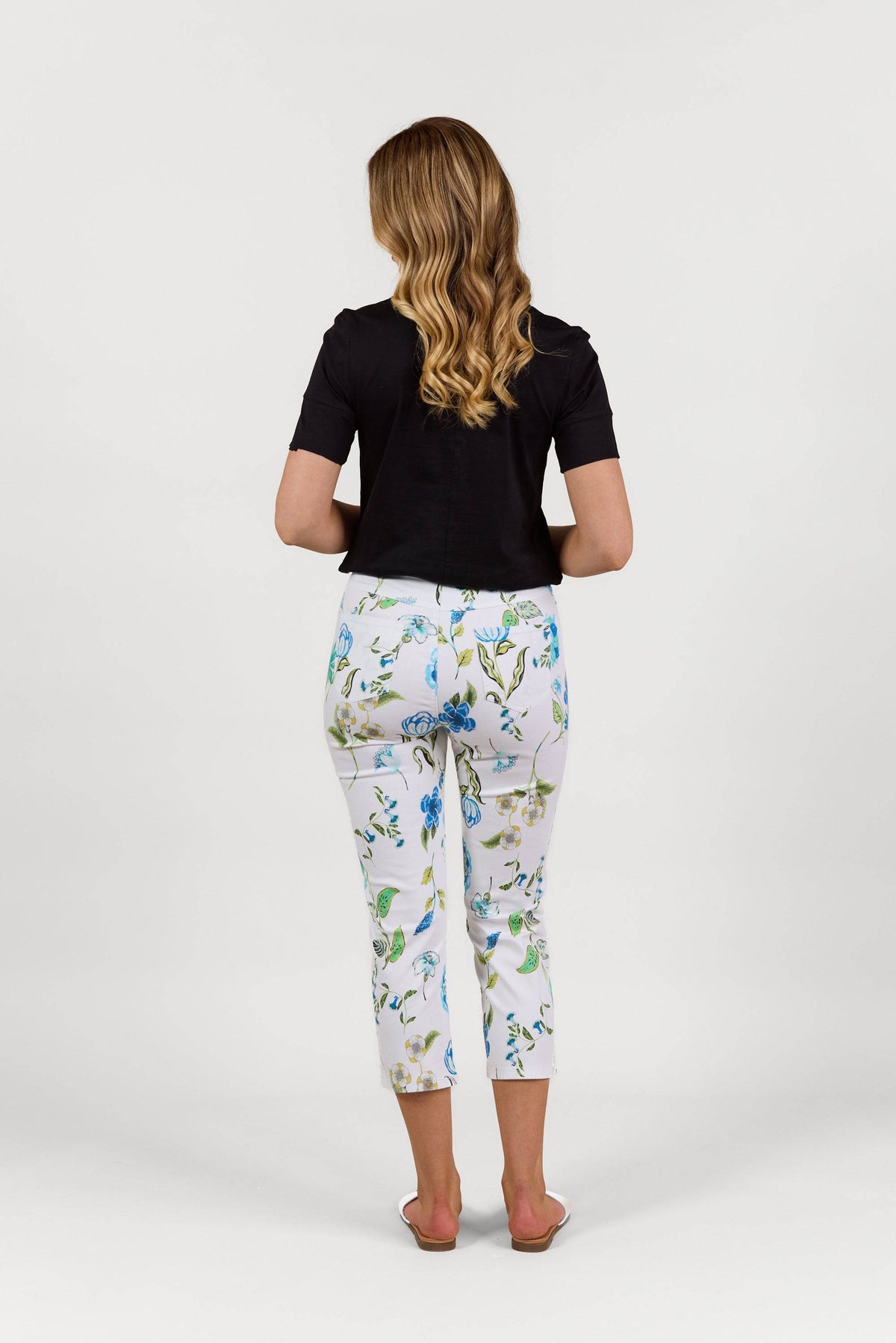 A woman with wavy blond hair wears a black short-sleeve top and Vassalli Sierra 7/8 Pant - Santorini, standing in white sandals facing away from the camera against a plain white background.