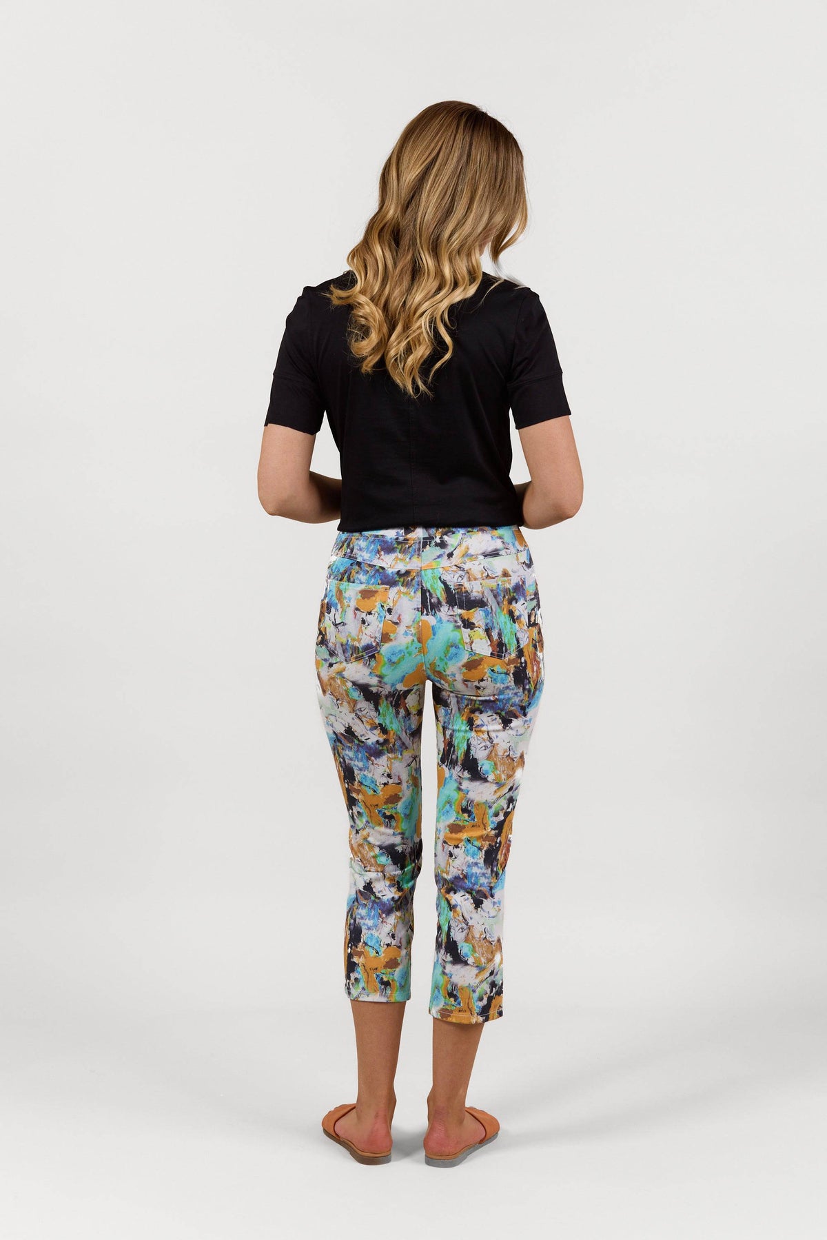 A woman with wavy blonde hair, seen from behind, wears a black short-sleeve top and Vassalli Sierra 7/8 Pant - Storm featuring colorful abstract prints, plus flat sandals, standing against a plain white background.