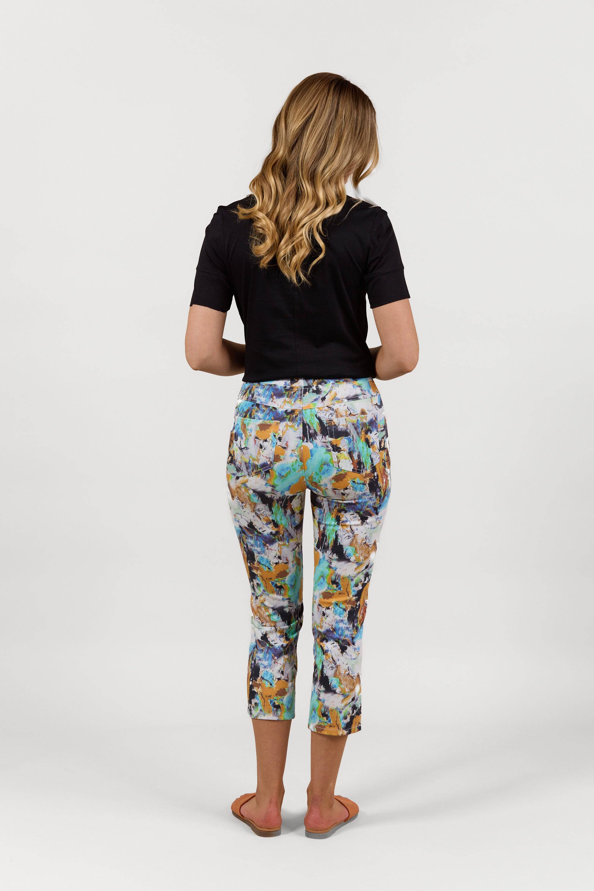 A woman with wavy blonde hair, seen from behind, wears a black short-sleeve top and Vassalli Sierra 7/8 Pant - Storm featuring colorful abstract prints, plus flat sandals, standing against a plain white background.