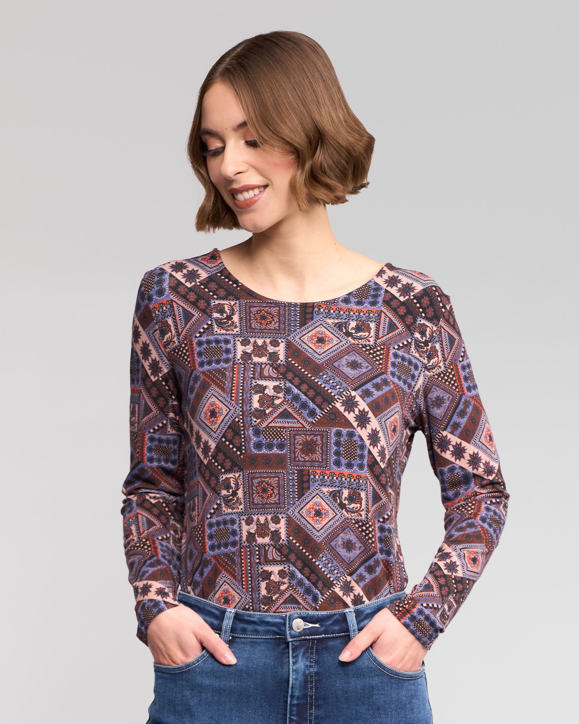 A woman with short brown hair wears the Vassalli Staple Top - Rue, a long-sleeve round-neck stretch jersey in blue, purple, and pink, paired with blue jeans. She smiles downwards with hands in her pockets against a light gray background.