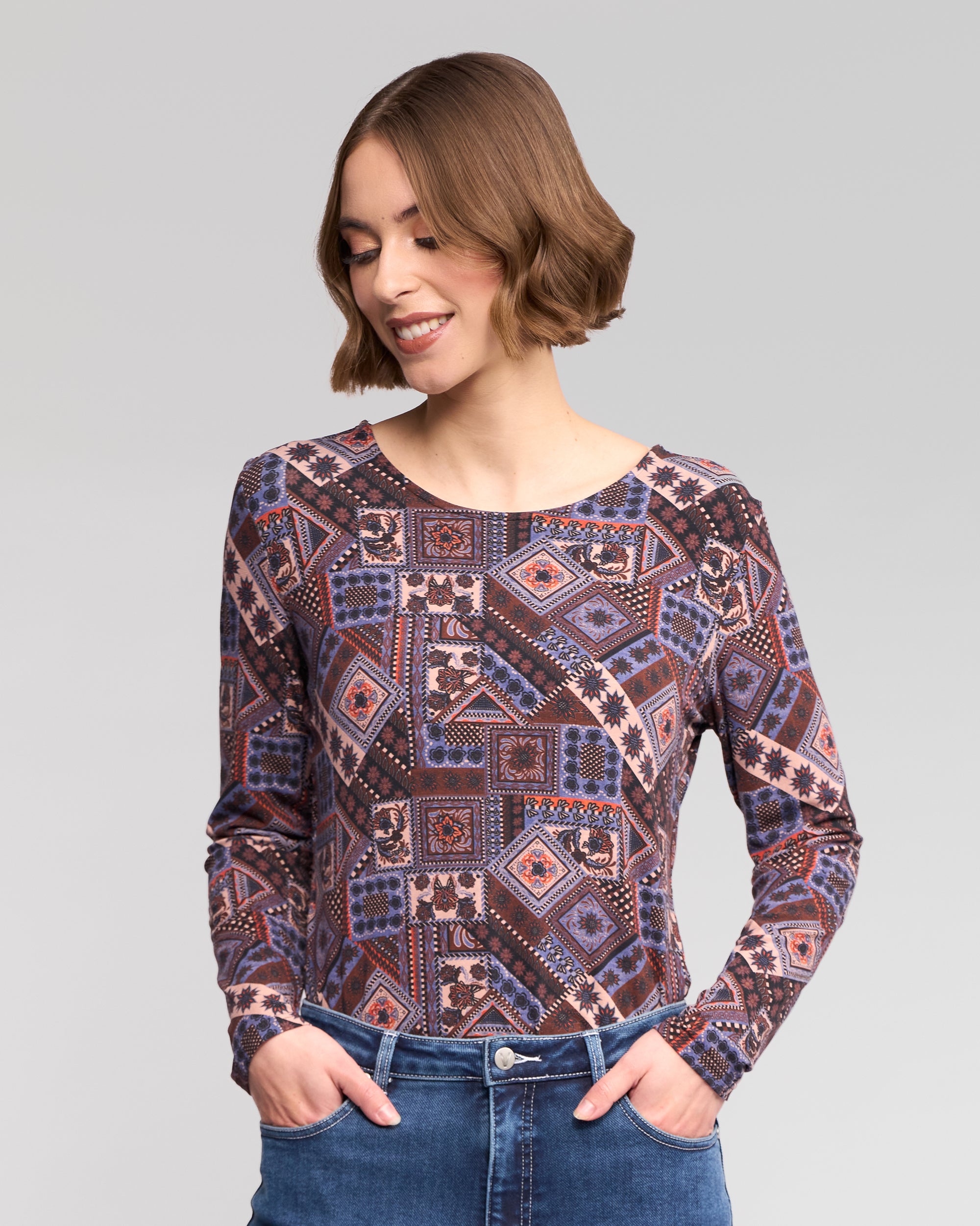 A woman with short brown hair wears the Vassalli Staple Top - Rue, a long-sleeve round-neck stretch jersey in blue, purple, and pink, paired with blue jeans. She smiles downwards with hands in her pockets against a light gray background.