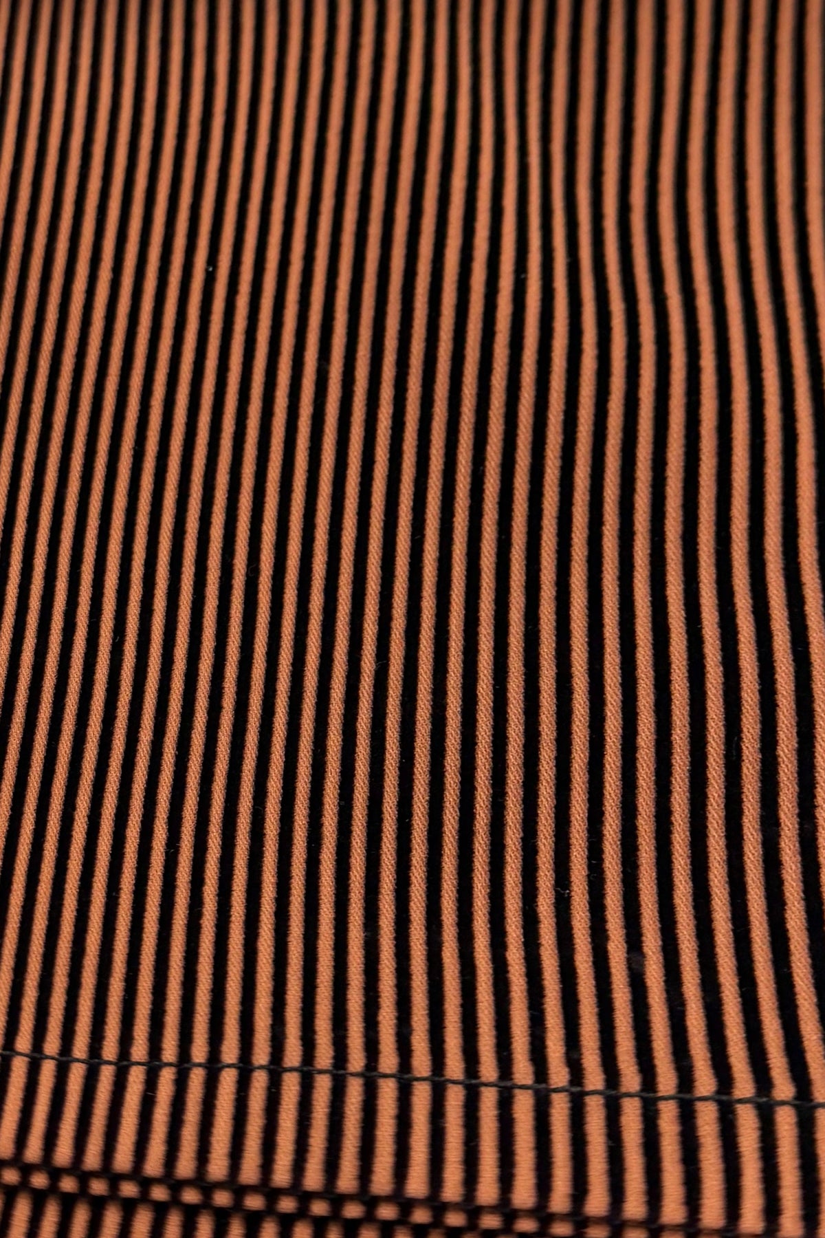 Close-up of the Bay Pull On - Tiger/Black Stripe by Vassalli, featuring stretch fabric with narrow vertical orange and black stripes that create an optical illusion effect. The woven texture and pattern evoke the look and feel of slim leg pull-on pants.