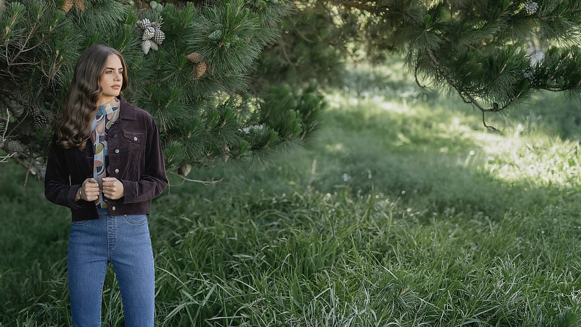 A woman in a brown jacket and blue jeans stands in tall grass near a pine tree, looking to the side. Sunlight filters through the branches, casting a natural, serene atmosphere.