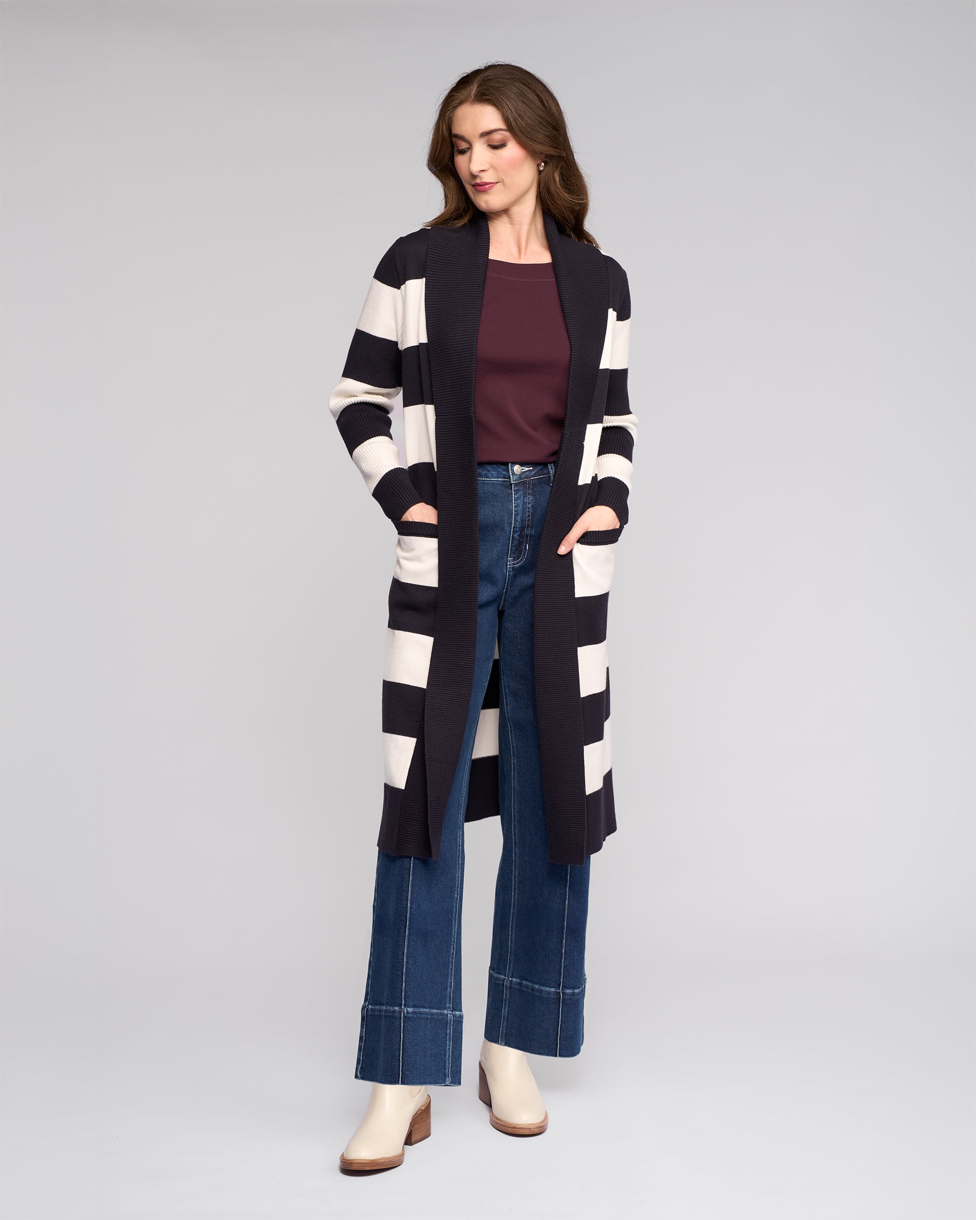 A woman poses against a plain background wearing the Vassalli Downtown Cardi in Ink/Winter White over a maroon top, paired with wide-leg blue jeans and light ankle boots, her hands tucked into the cardigan pockets.