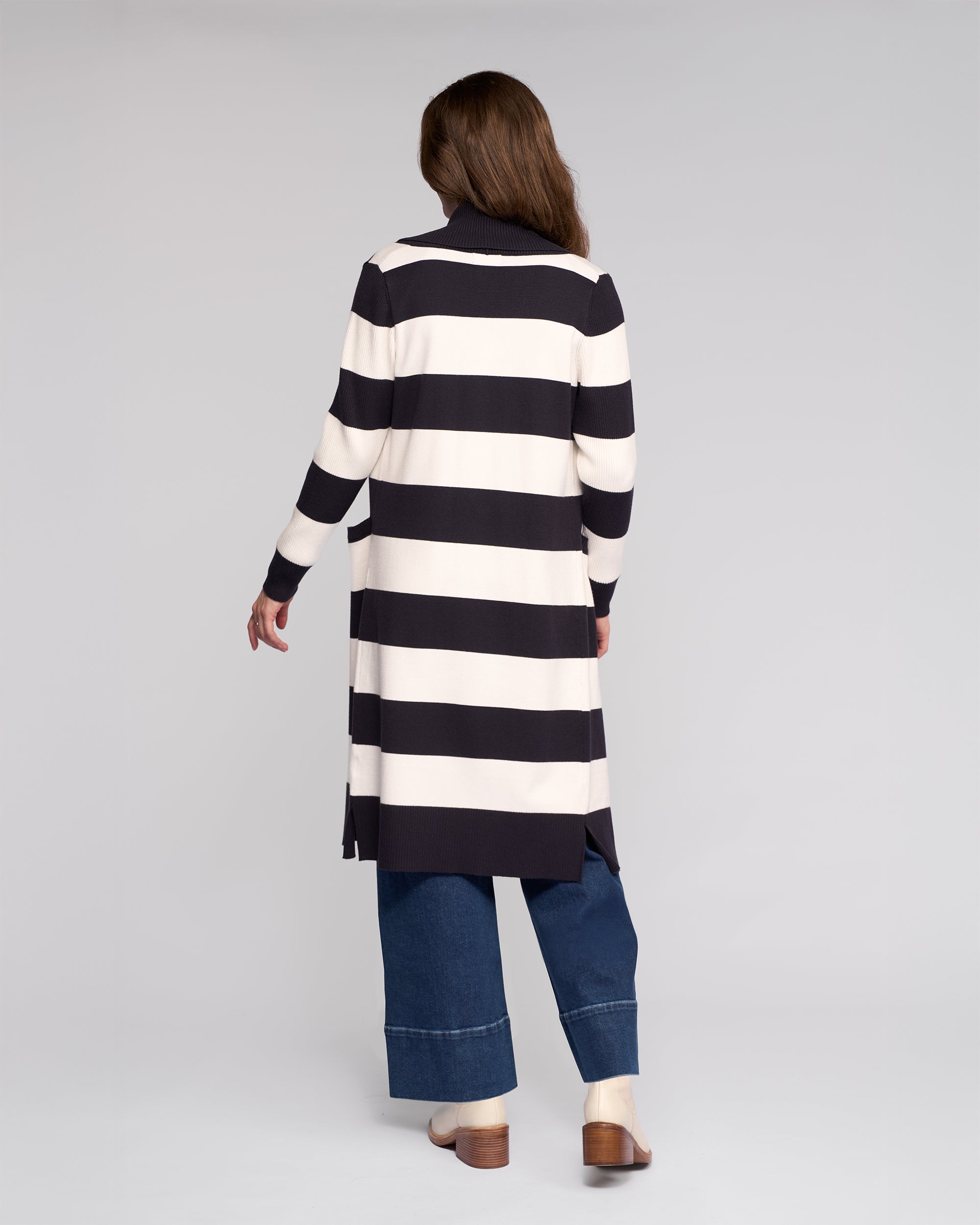 A woman faces away, wearing the Vassalli Downtown Cardi in Ink/Winter White with wide-leg blue jeans and cream ankle boots, standing against a plain light gray background.