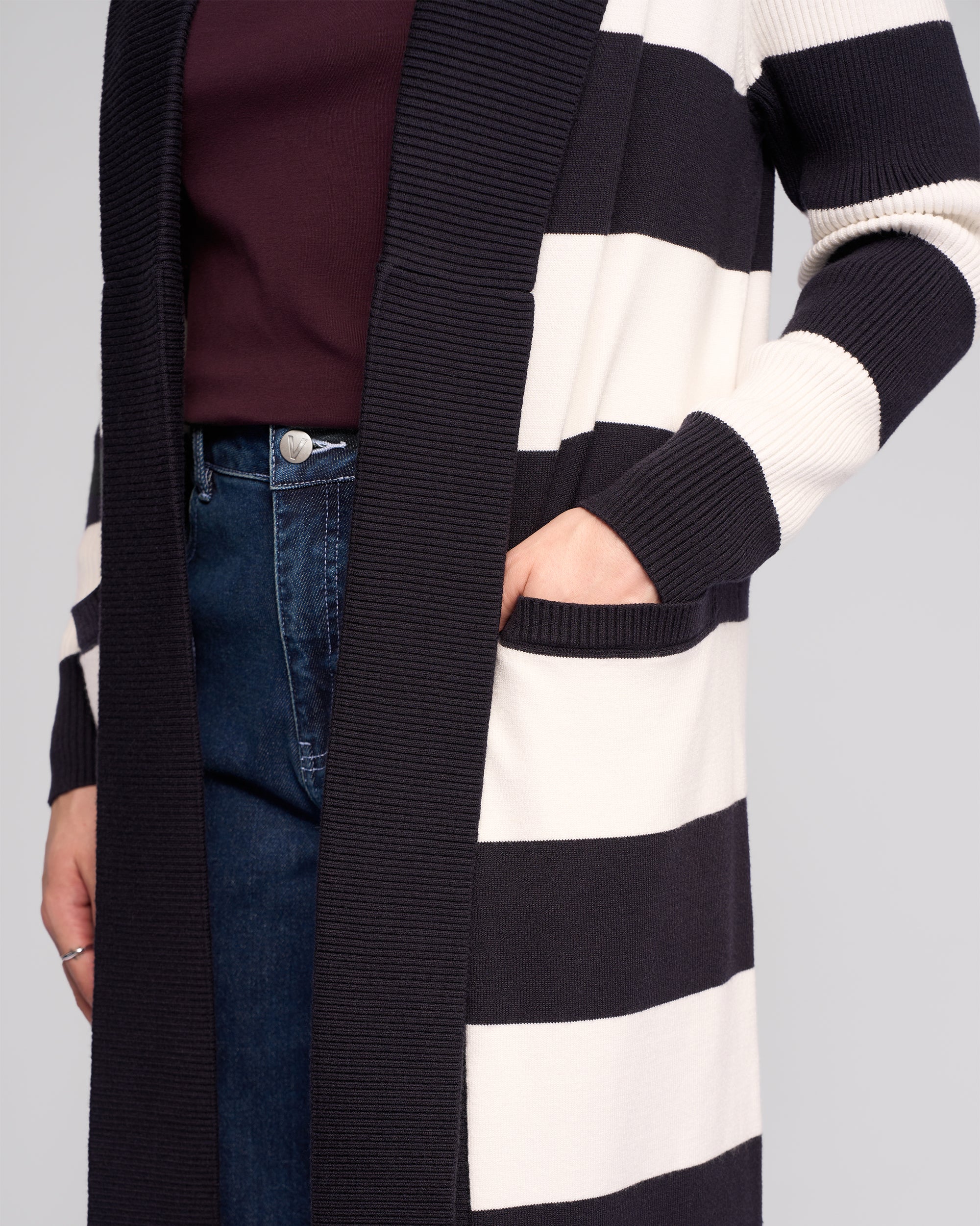 A person wears a maroon top, dark blue jeans, and the Vassalli Downtown Cardi - Ink/Winter White, styled open with one hand in the pocket. The image shows the relaxed-fit cardigan from shoulders to mid-thigh.