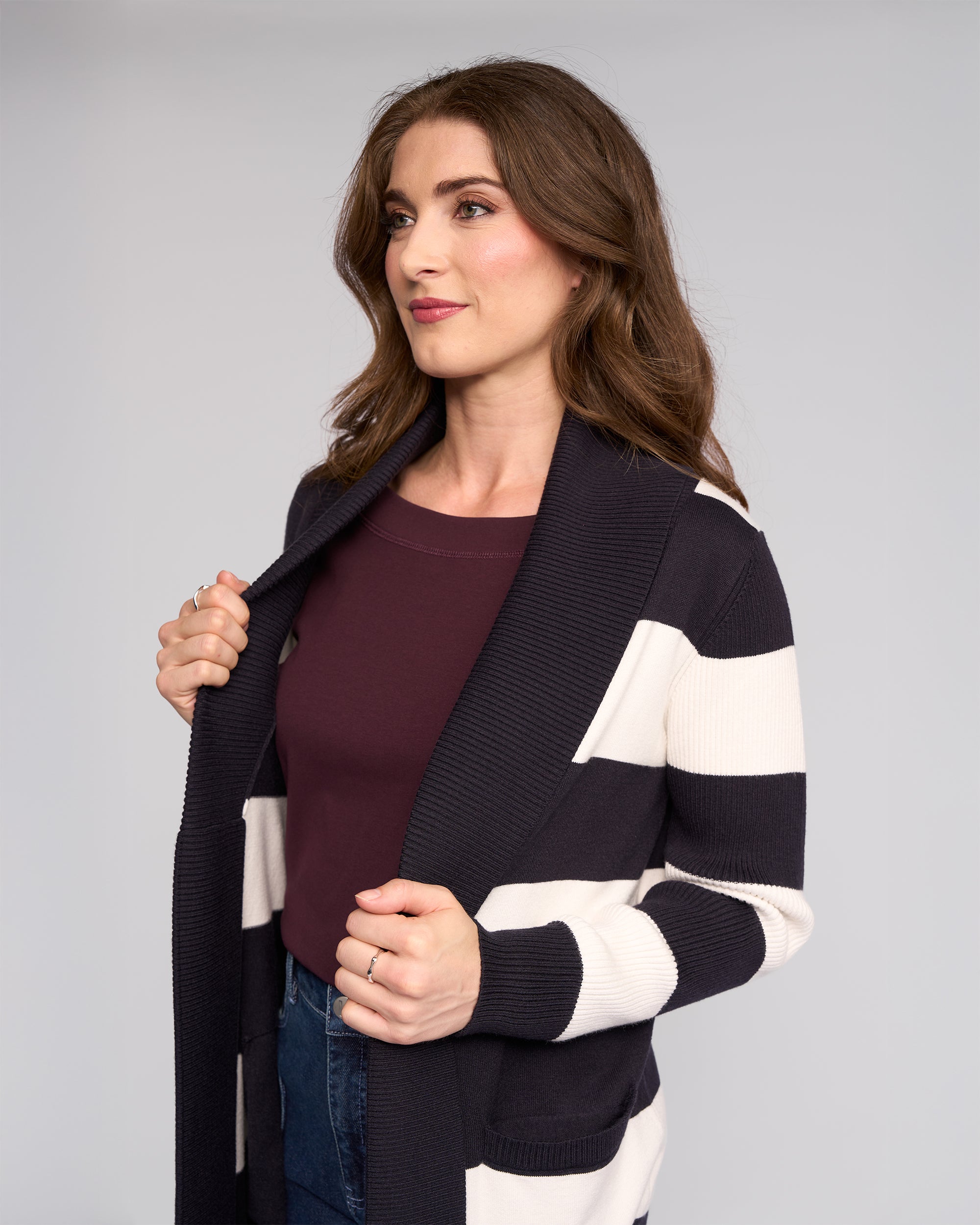 A woman with wavy brown hair, in a maroon top and the Vassalli Downtown Cardi - Ink/Winter White, looks slightly to the side with a soft smile while holding the edges of her cardigan.