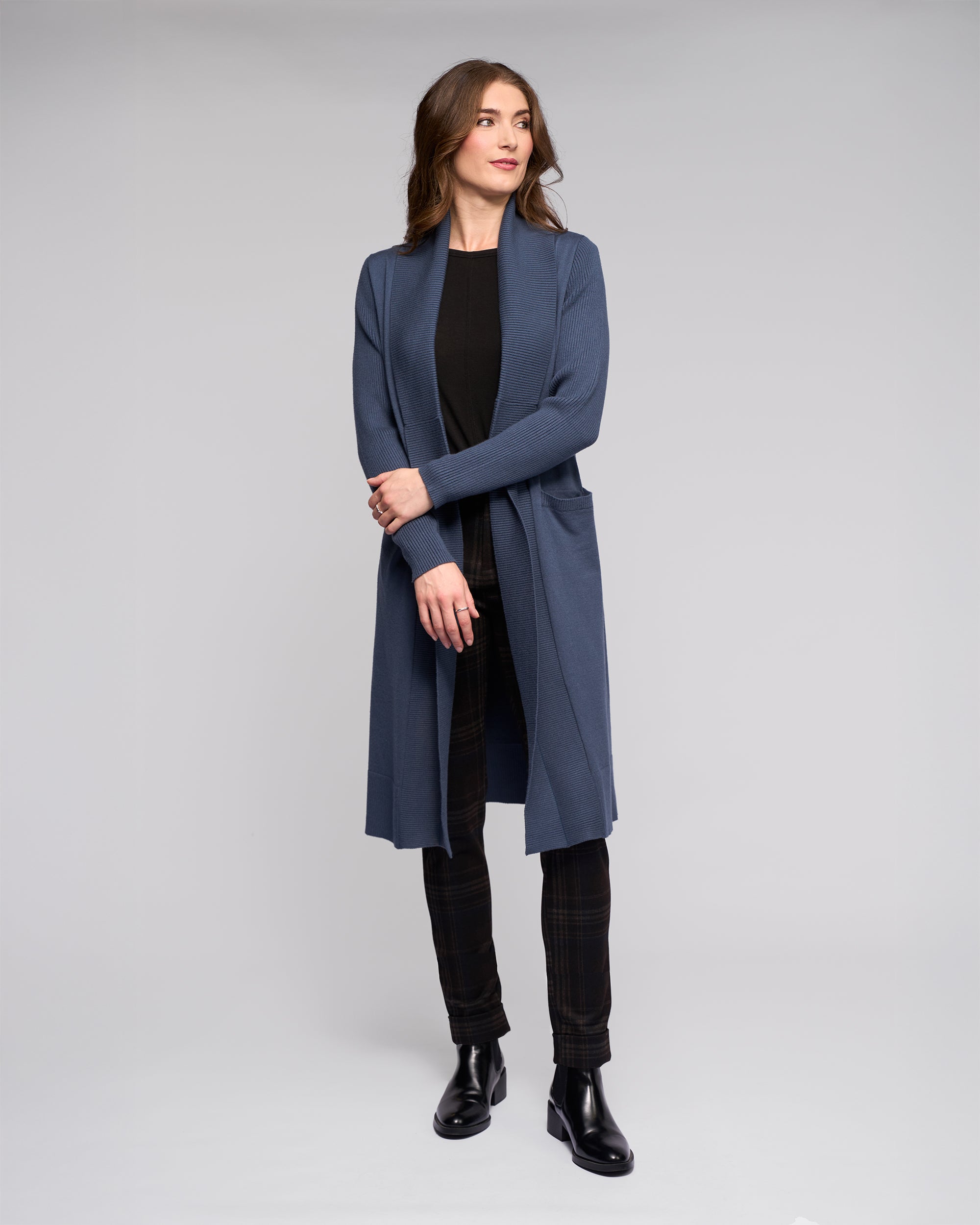 A woman stands against a plain background wearing Vassalli’s Downtown Cardi in Marine over a black top, dark plaid pants, and black ankle boots. She gazes to the side, gently clasping her hands.
