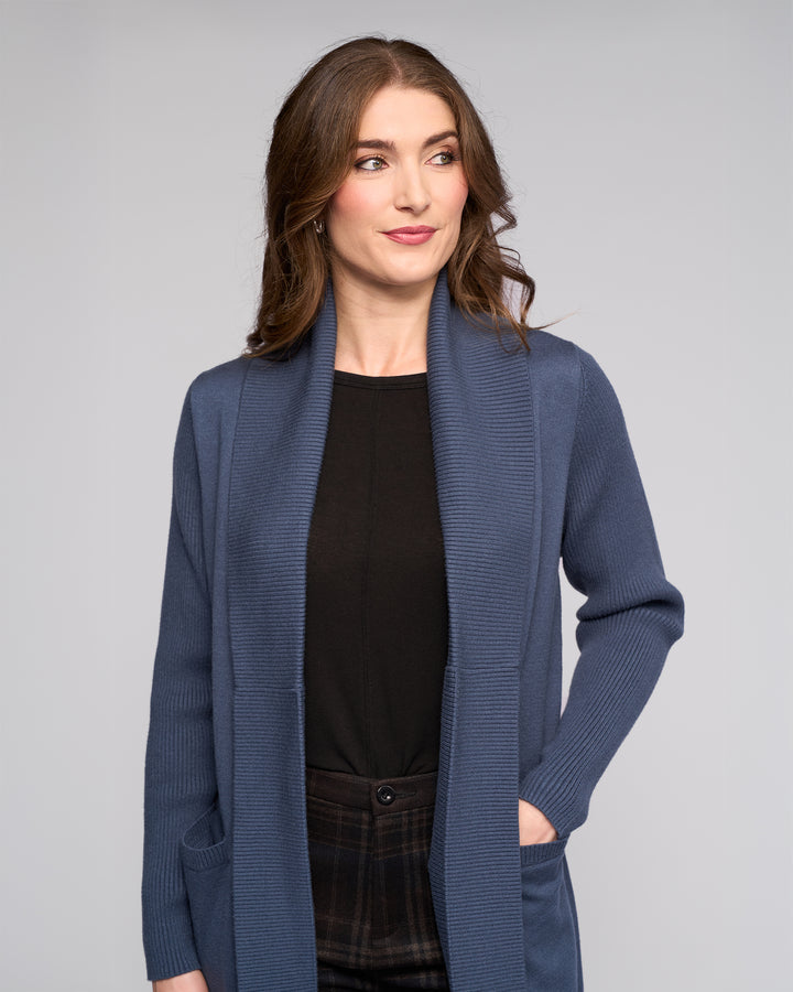 A woman with wavy brown hair wears the Vassalli Downtown Cardi in Marine over a black top and plaid pants, standing with one hand in her pocket and looking slightly to the side against a plain light gray background.