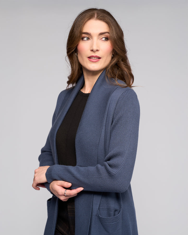 A woman with long brown hair wears the Vassalli Downtown Cardi in Marine over a black top, standing against a light gray background. She looks to her left with a slight smile, arms loosely crossed.