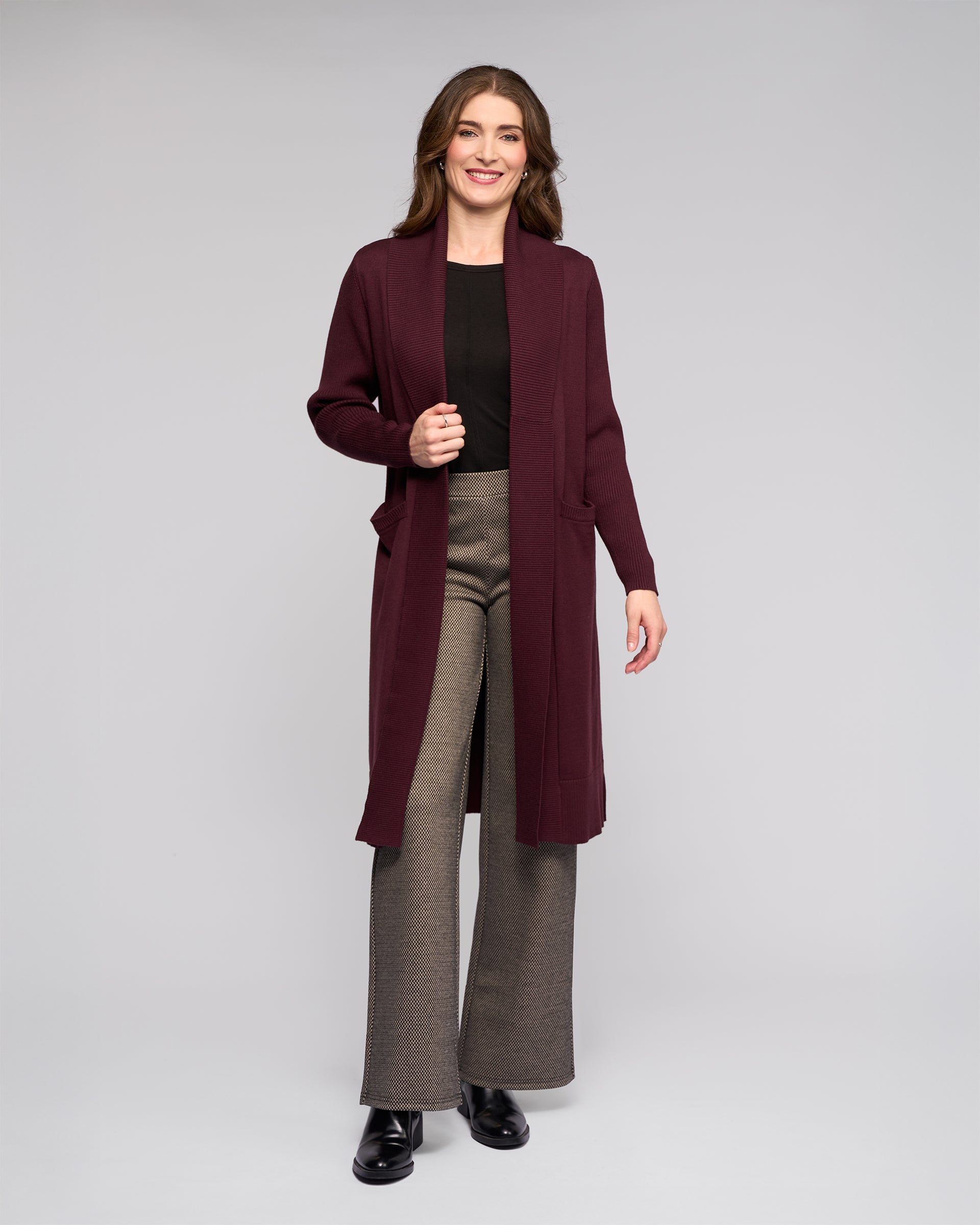 A woman with long brown hair smiles while wearing the Vassalli Downtown Cardi in Merlot over a black top, paired with gray wide-leg pants and black shoes, standing against a plain light gray background.