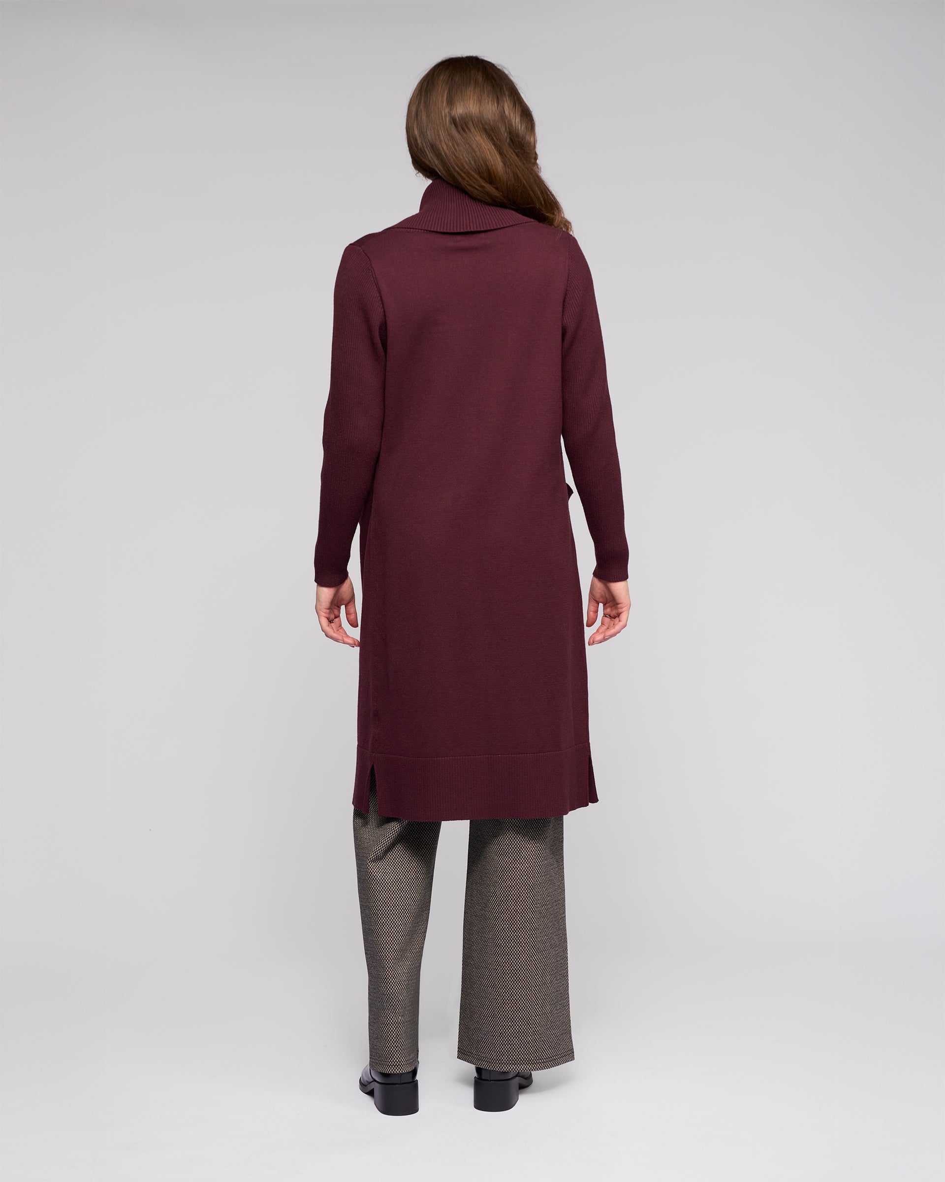 A woman, turned away, wears the Vassalli Downtown Cardi in Merlot over a turtleneck with gray pants and black shoes, set against a plain light gray background.