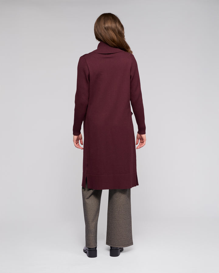A woman, turned away, wears the Vassalli Downtown Cardi in Merlot over a turtleneck with gray pants and black shoes, set against a plain light gray background.