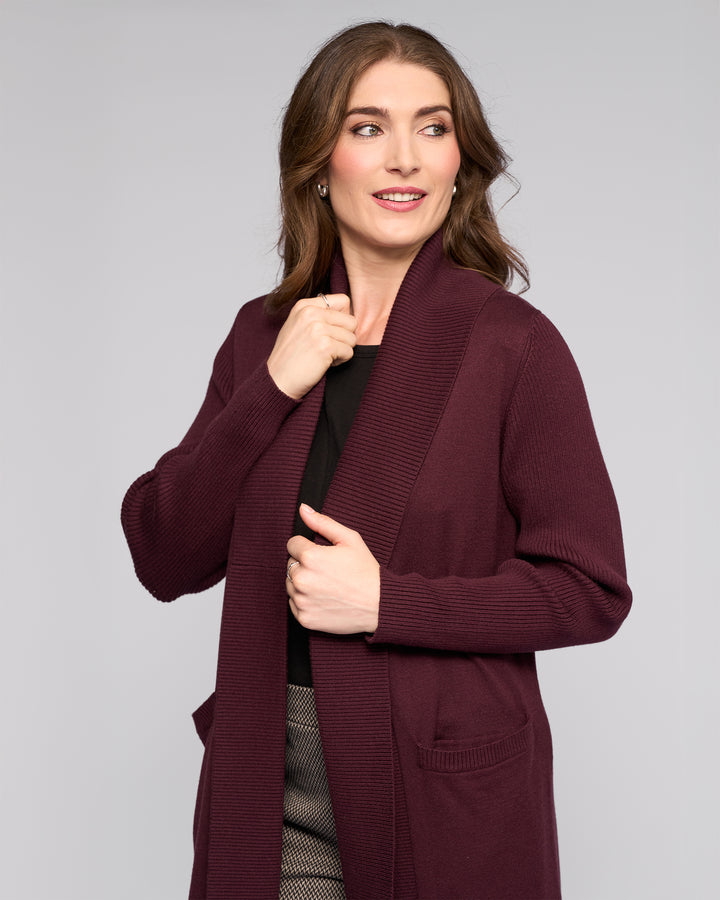 A woman wears the Vassalli Downtown Cardi in Merlot, featuring ribbed trim and patch pockets, layered over a black top and patterned pants, smiling against a grey background.