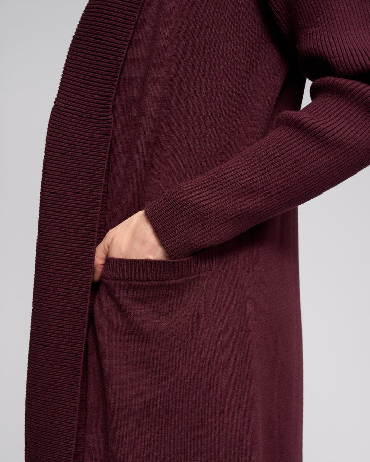 A person wears the Vassalli Downtown Cardi in Merlot, featuring long sleeves and ribbed detailing, while placing their right hand into a side pocket. The background is plain and light-colored.