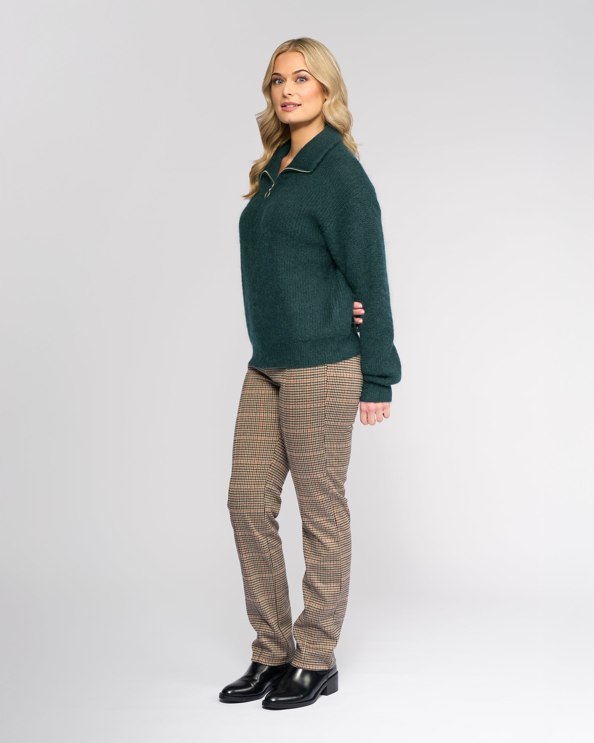 A woman with long blonde hair stands slightly turned, wearing the Vassalli Origin Sweater in Mallard with plaid pants and black shoes, set against a plain light gray background.