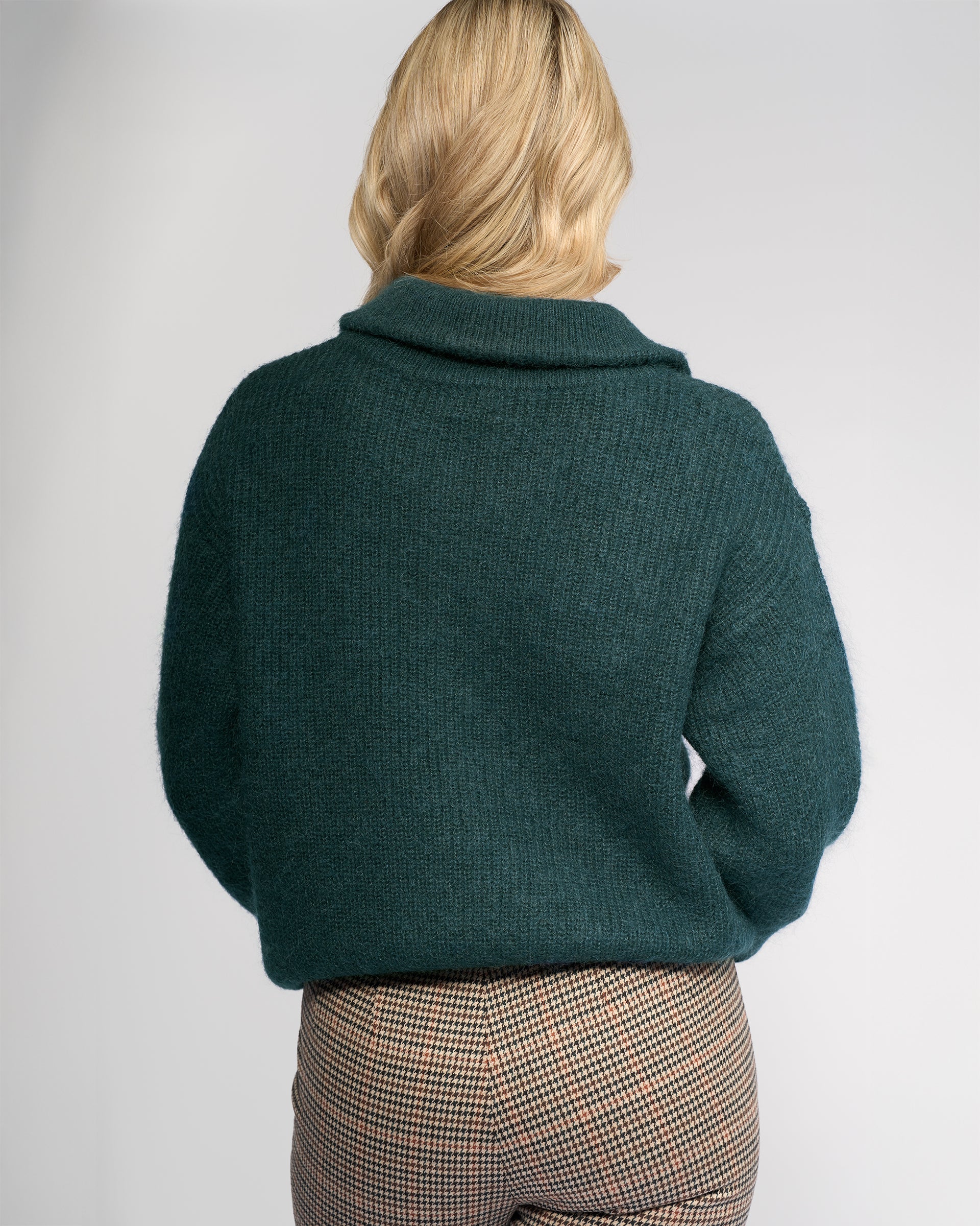 A person with blonde, shoulder-length hair stands with their back to the camera, wearing the Vassalli Origin Sweater in Mallard and brown plaid pants against a plain light background.