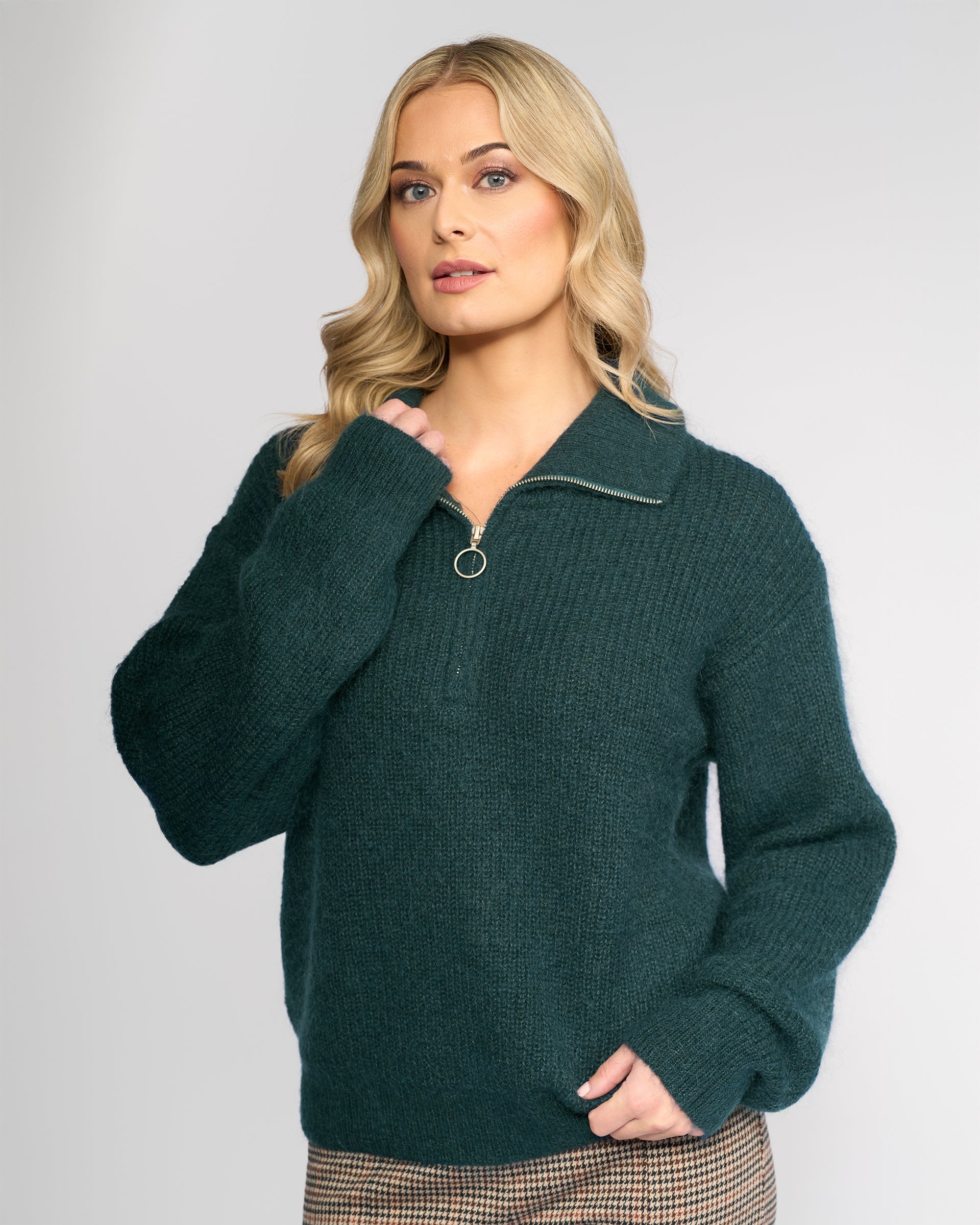 A woman with long blonde hair models the Vassalli Origin Sweater in Mallard, a chunky rib knit dark green half zip, paired with plaid pants. She stands confidently against a plain light background, one hand near her chest and the other in her pocket.
