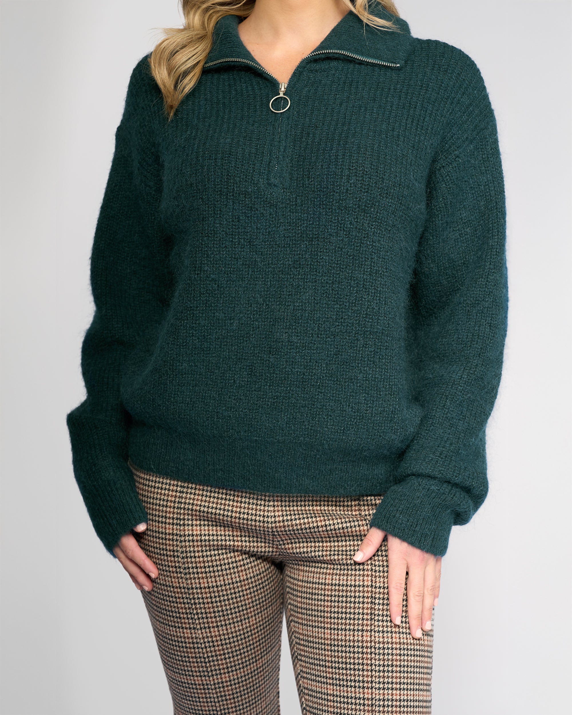 A person wears the Vassalli Origin Sweater - Mallard, a dark green chunky rib knit half zip with ring pull, paired with brown plaid pants and stands against a plain light background. The person’s head is partially out of frame.
