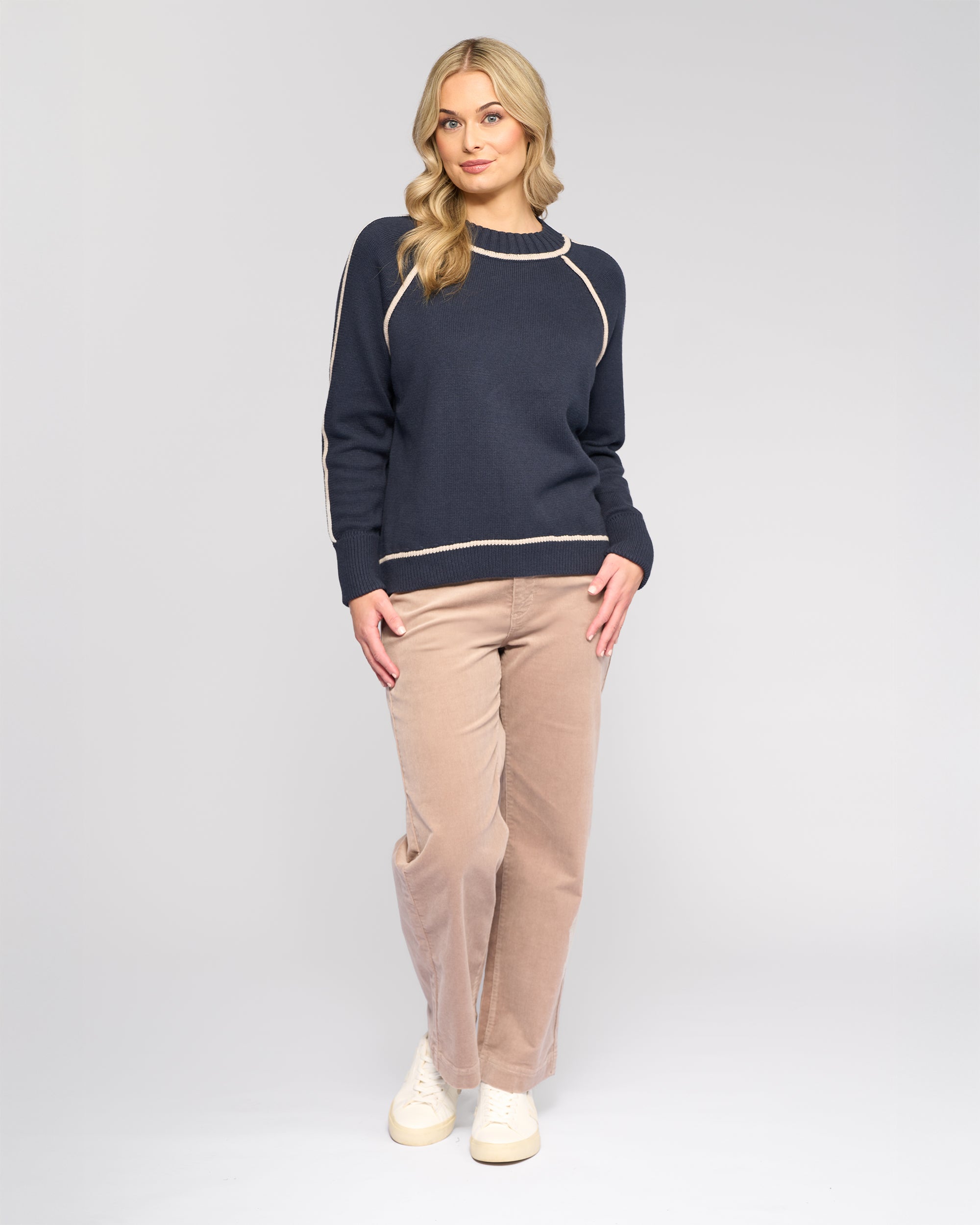 A woman with long blonde hair wears the Vassalli Zane Sweater in Ink/Oat with beige pants and white sneakers, standing against a plain light gray background and looking at the camera with a slight smile.