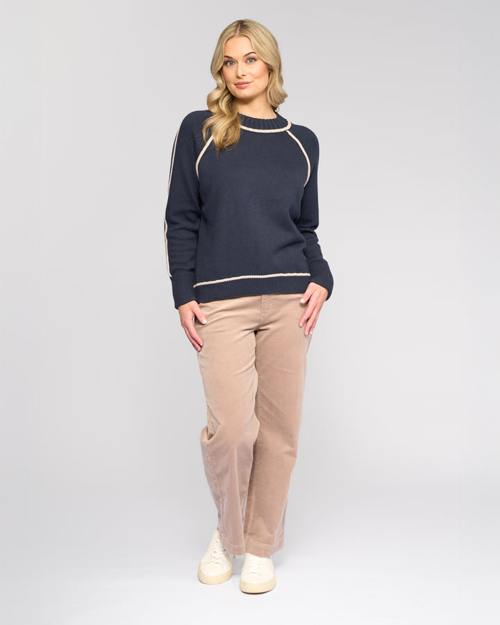 A woman with long blonde hair wears the Vassalli Zane Sweater in Ink/Oat with beige pants and white sneakers, standing against a plain light gray background and looking at the camera with a slight smile.