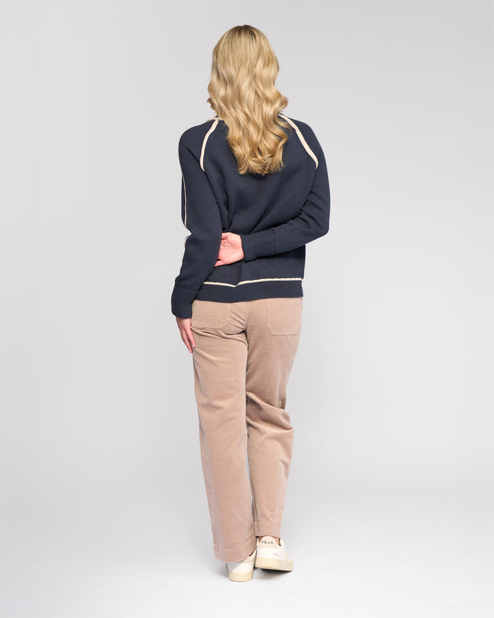 A person with long, wavy blonde hair stands facing away, wearing the Vassalli Zane Sweater in Ink/Oat, paired with beige pants and white shoes, one hand behind their back against a plain light background.