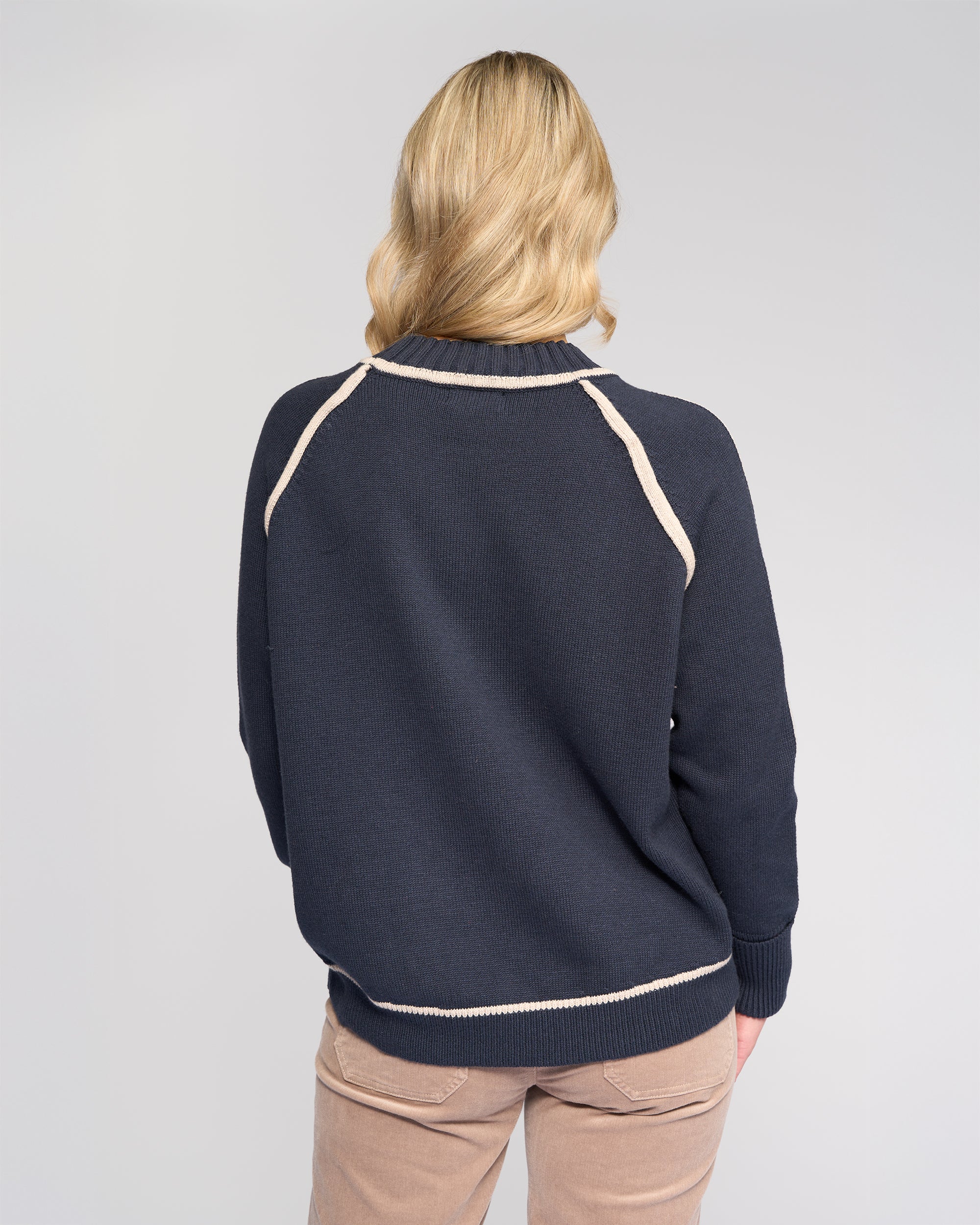 A person with blonde, wavy hair stands with their back to the camera, wearing the Vassalli Zane Sweater in Ink/Oat and beige pants, set against a plain, light background.