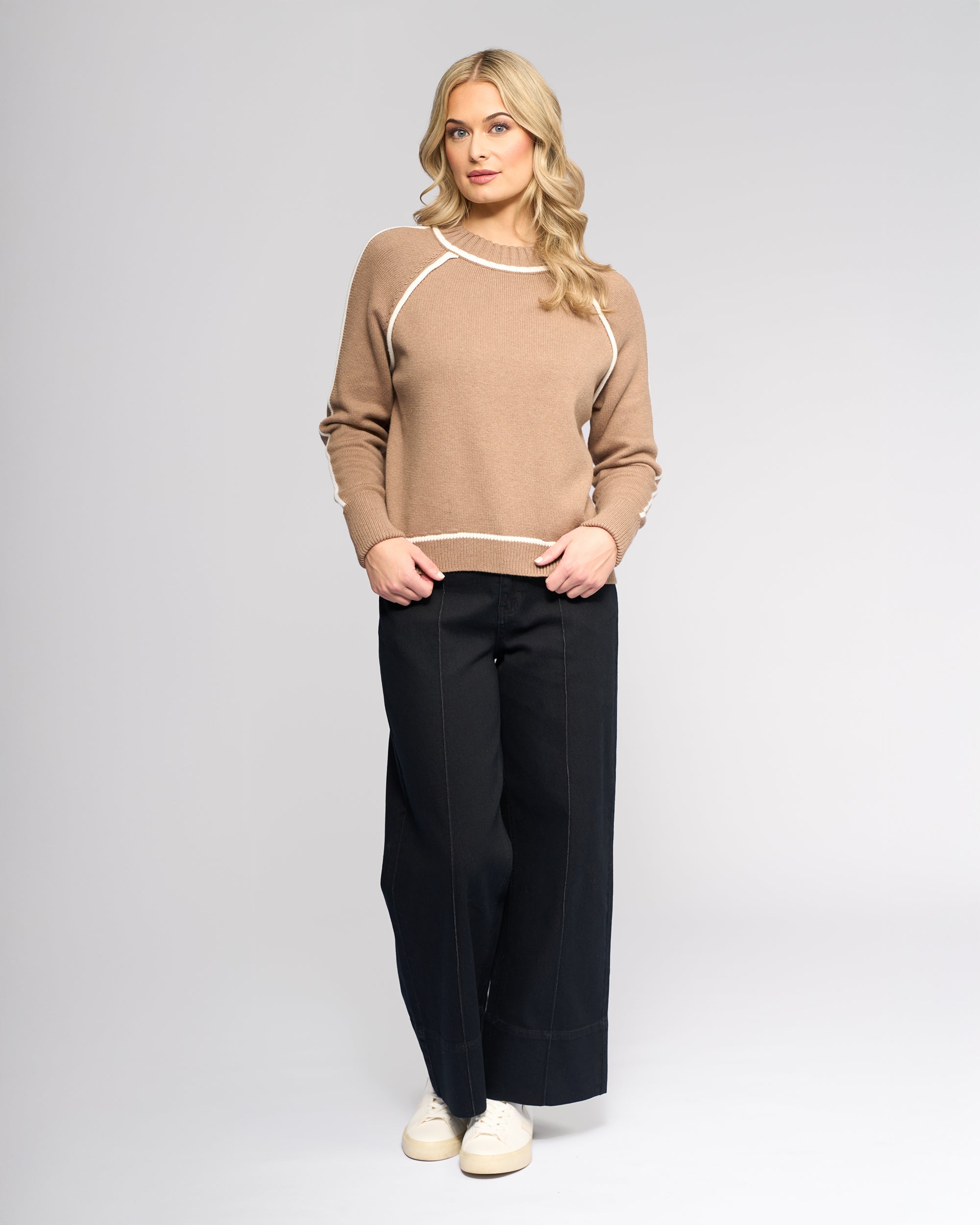 A woman with long blonde hair wears the Vassalli Zane Sweater in Twig/Cream, wide-leg black pants with white stitching, and white sneakers, standing against a plain light gray background.