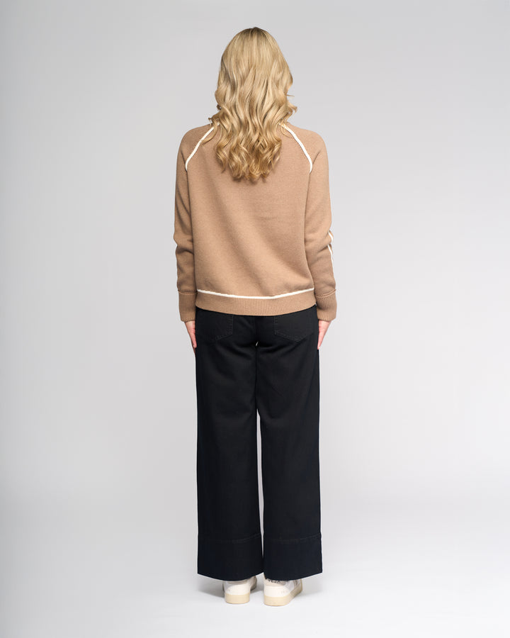 A person with long, wavy blonde hair stands facing away, wearing the Vassalli Zane Sweater in Twig/Cream, black wide-leg pants, and white shoes against a plain light gray background.