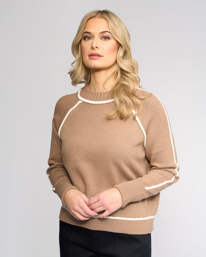A woman with long blonde hair and light makeup models the Vassalli Zane Sweater in Twig/Cream, paired with black pants. She stands against a plain light gray background, facing forward with hands loosely clasped.