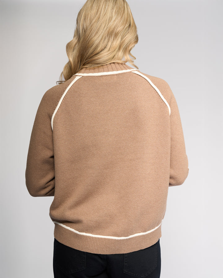 A person with long, wavy blonde hair is seen from behind, wearing the Vassalli Zane Sweater in Twig/Cream and dark pants, standing against a plain light background.