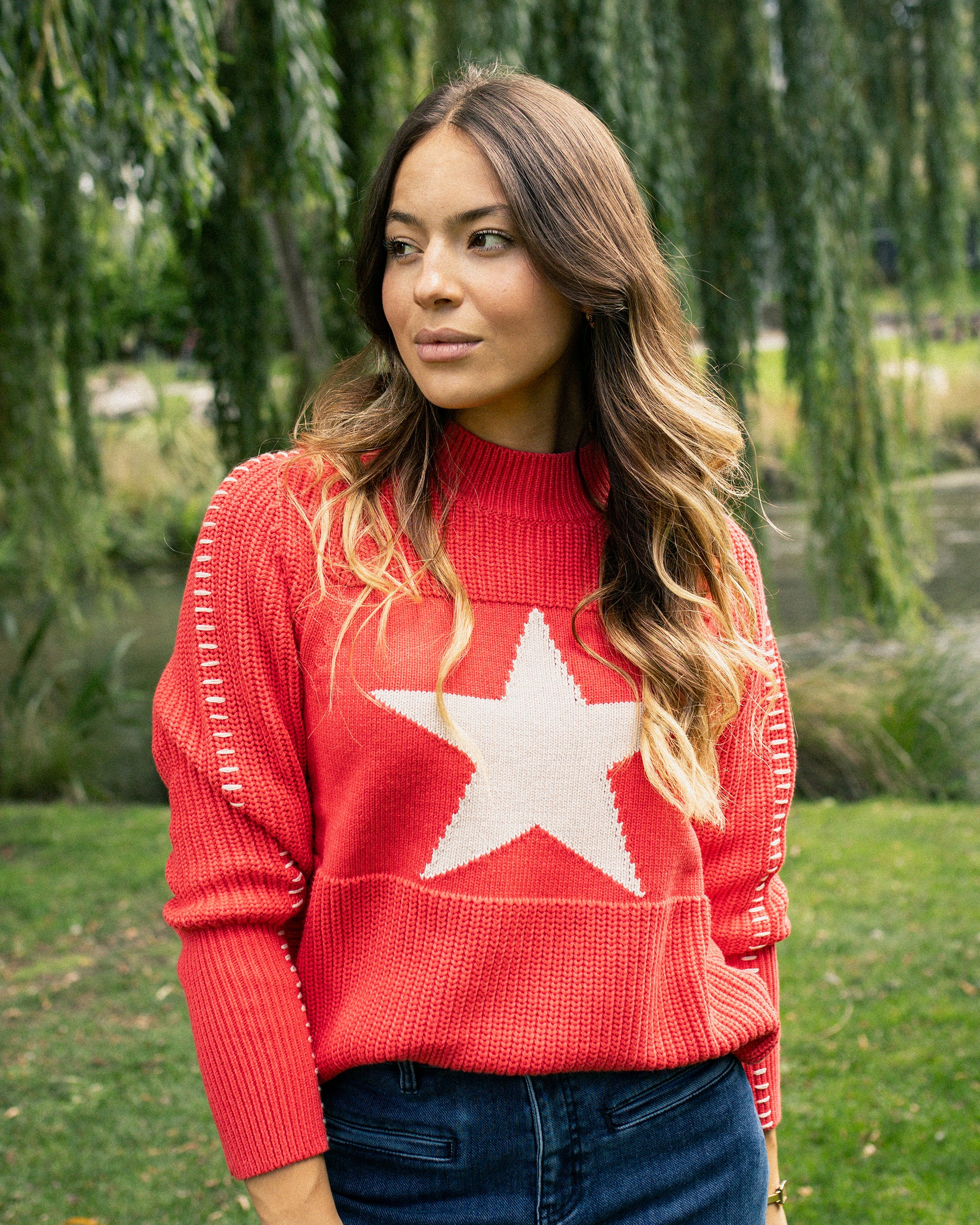A woman with long wavy hair stands outdoors by trees, wearing the Vassalli Star Sweater in coral with a bold white star on the front and blue jeans, gazing to the side with a thoughtful look.