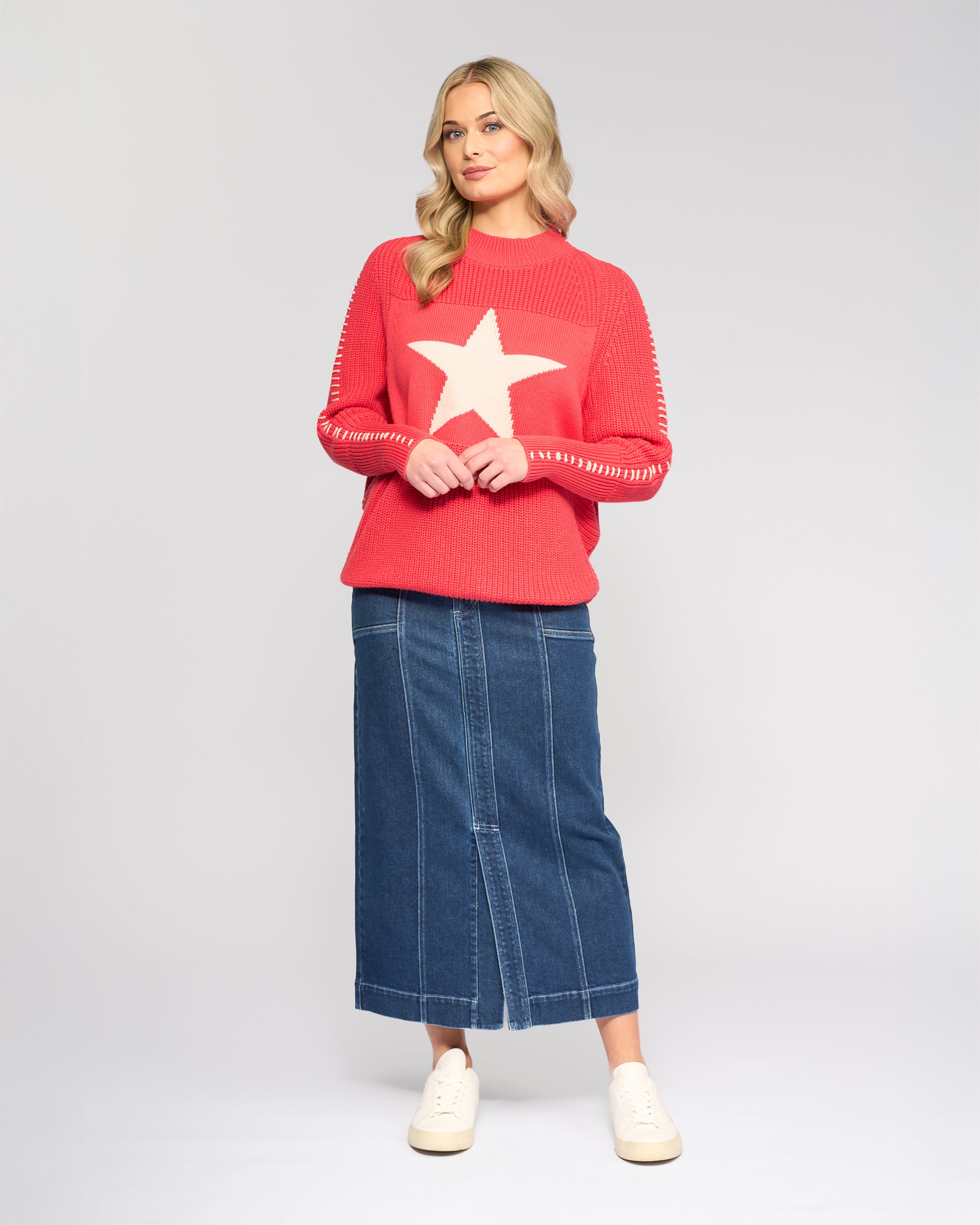 A woman with blonde wavy hair stands against a light gray background, wearing the Vassalli Star Sweater in coral, featuring a large white star on the front, paired with a long blue denim skirt and white sneakers.