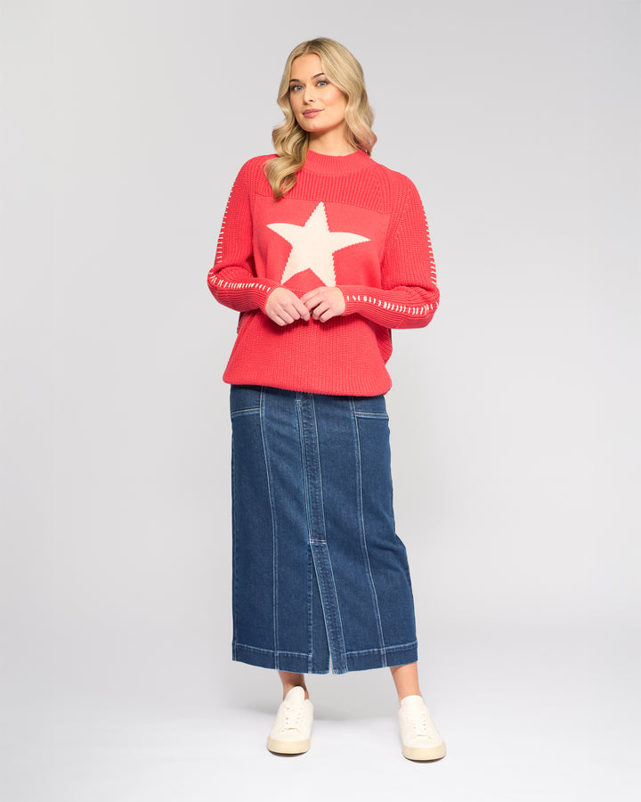 A woman with blonde wavy hair stands against a light gray background, wearing the Vassalli Star Sweater in coral, featuring a large white star on the front, paired with a long blue denim skirt and white sneakers.