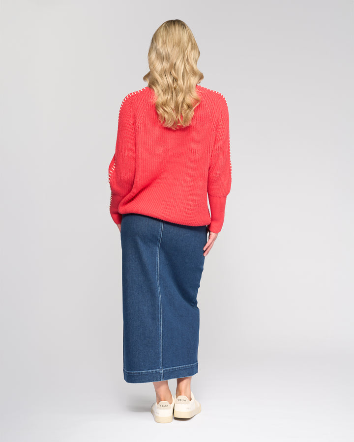A woman with long wavy blonde hair stands facing away, wearing the Vassalli Star Sweater in Coral, a blue denim midi skirt, and cream slip-on shoes against a plain light background.