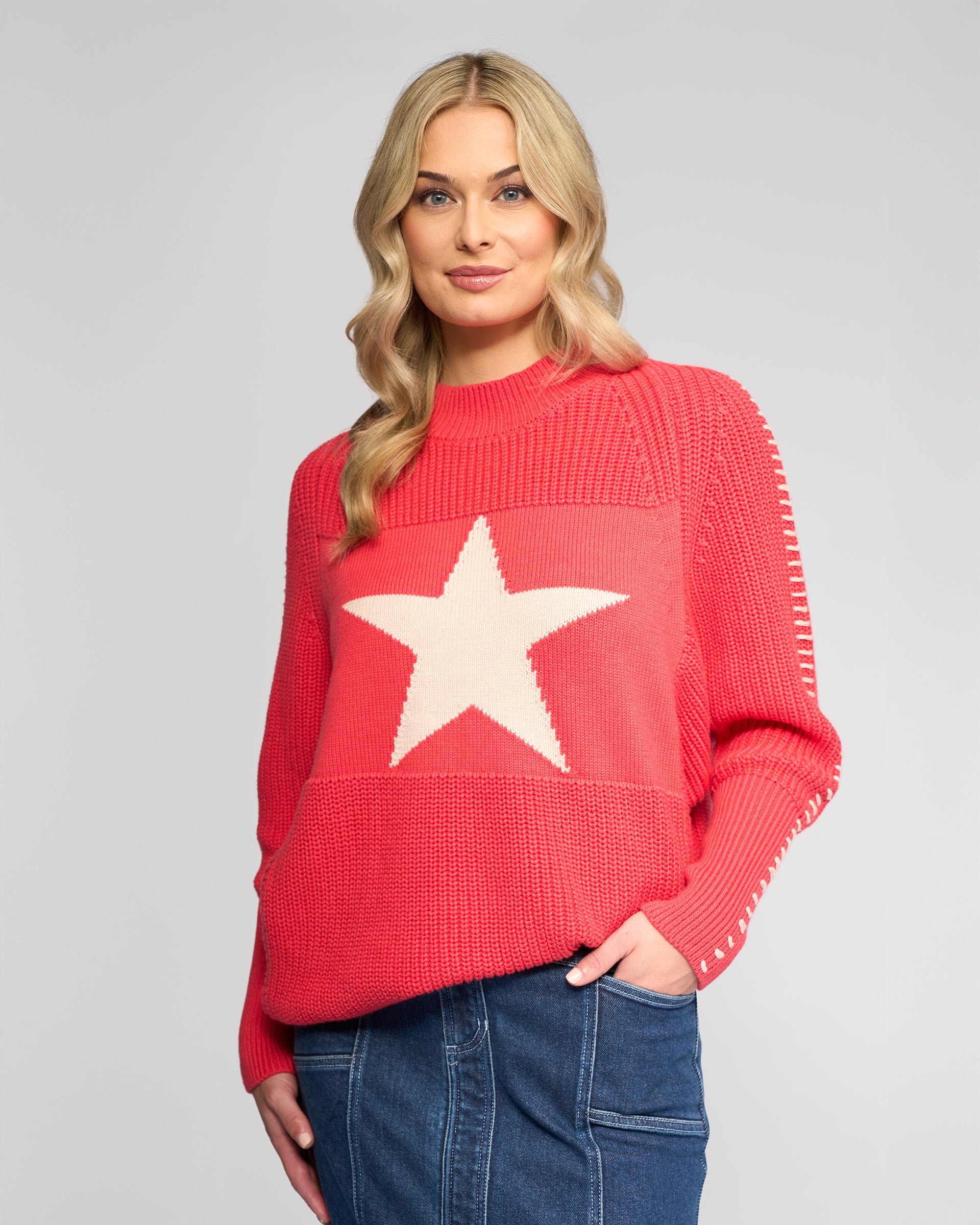 A woman with long blonde hair wears the Vassalli Star Sweater - Coral, an oversized rib-knit pink sweater with a large white star on the front, paired with a blue denim skirt against a plain light gray background.