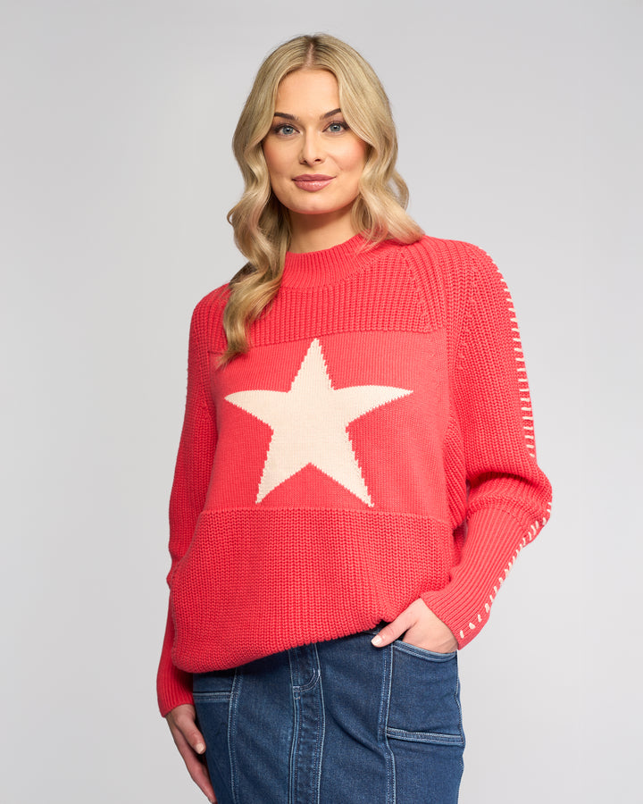 A woman with long blonde hair wears the Vassalli Star Sweater - Coral, an oversized rib-knit pink sweater with a large white star on the front, paired with a blue denim skirt against a plain light gray background.