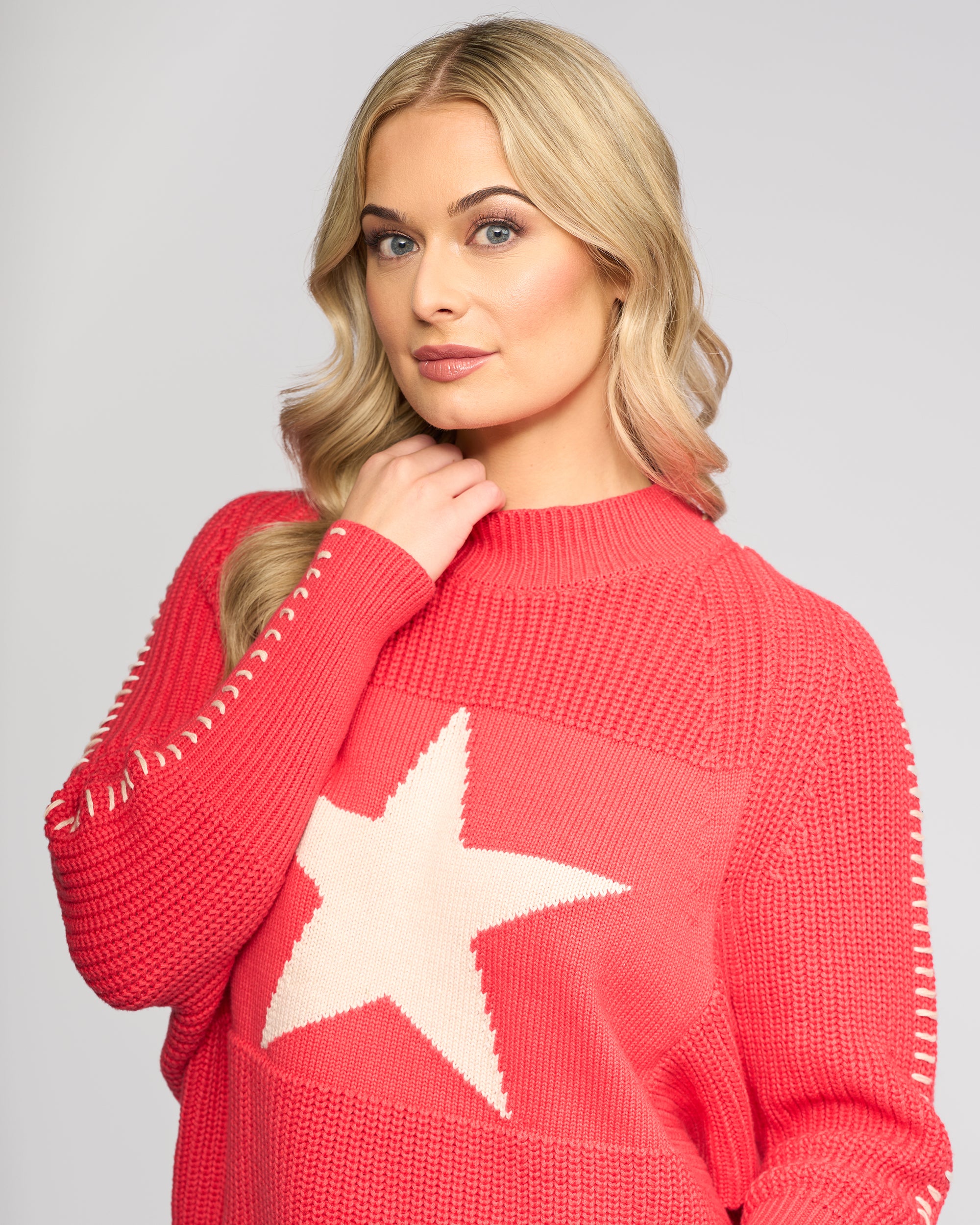 A woman with long blonde hair models the Vassalli Star Sweater - Coral, an oversized rib-knit style with a large white star on the front, as she poses confidently against a light gray background, gently holding the collar.