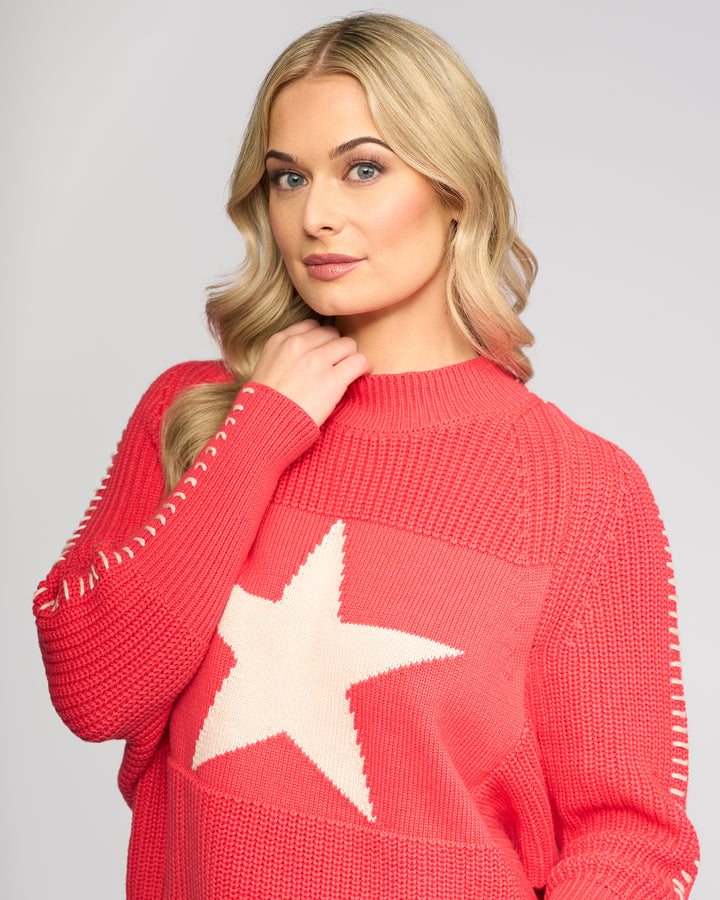 A woman with long blonde hair models the Vassalli Star Sweater - Coral, an oversized rib-knit style with a large white star on the front, as she poses confidently against a light gray background, gently holding the collar.