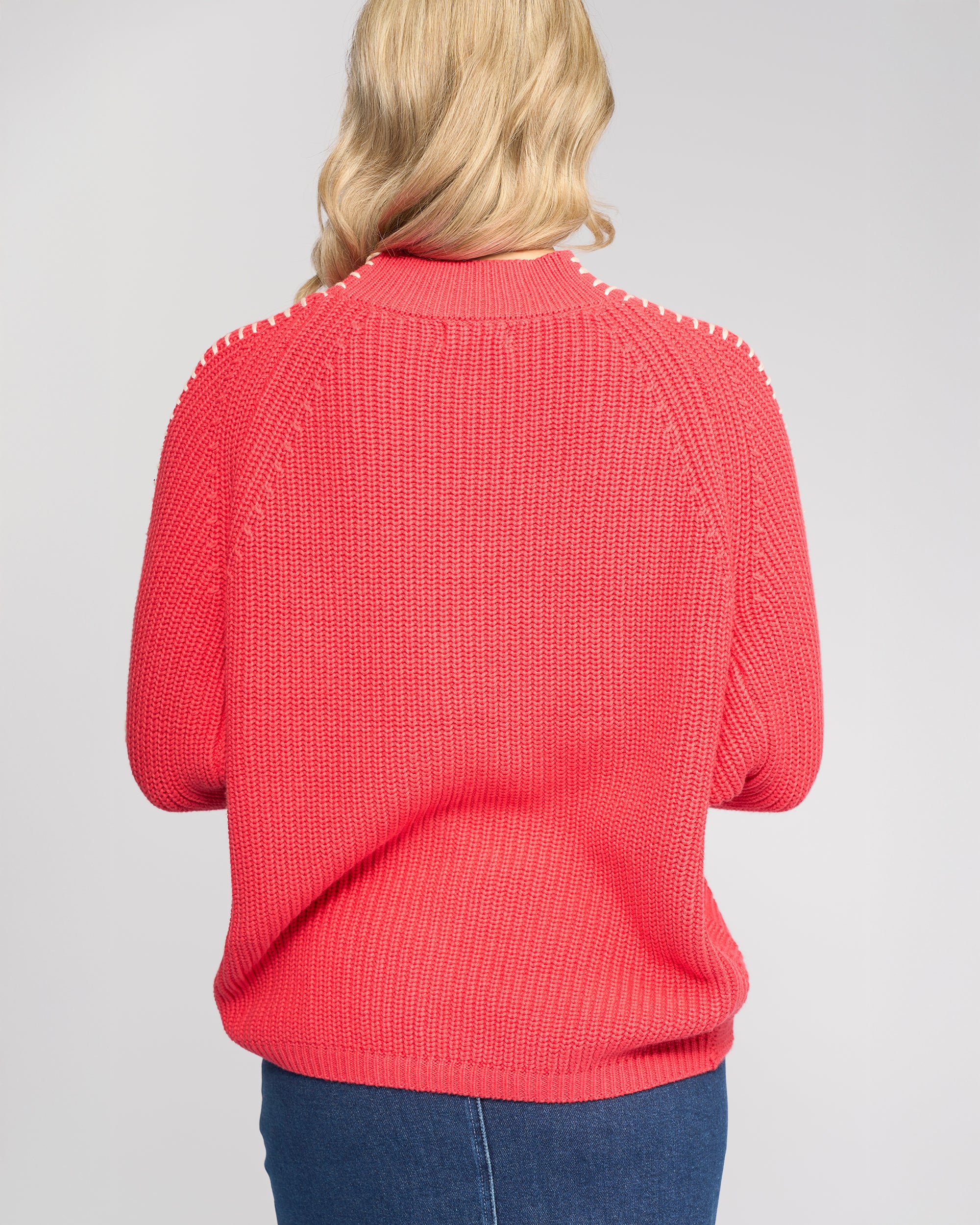 A person with long blonde hair, seen from behind, wears the Vassalli Star Sweater in coral and blue jeans against a plain light gray background.