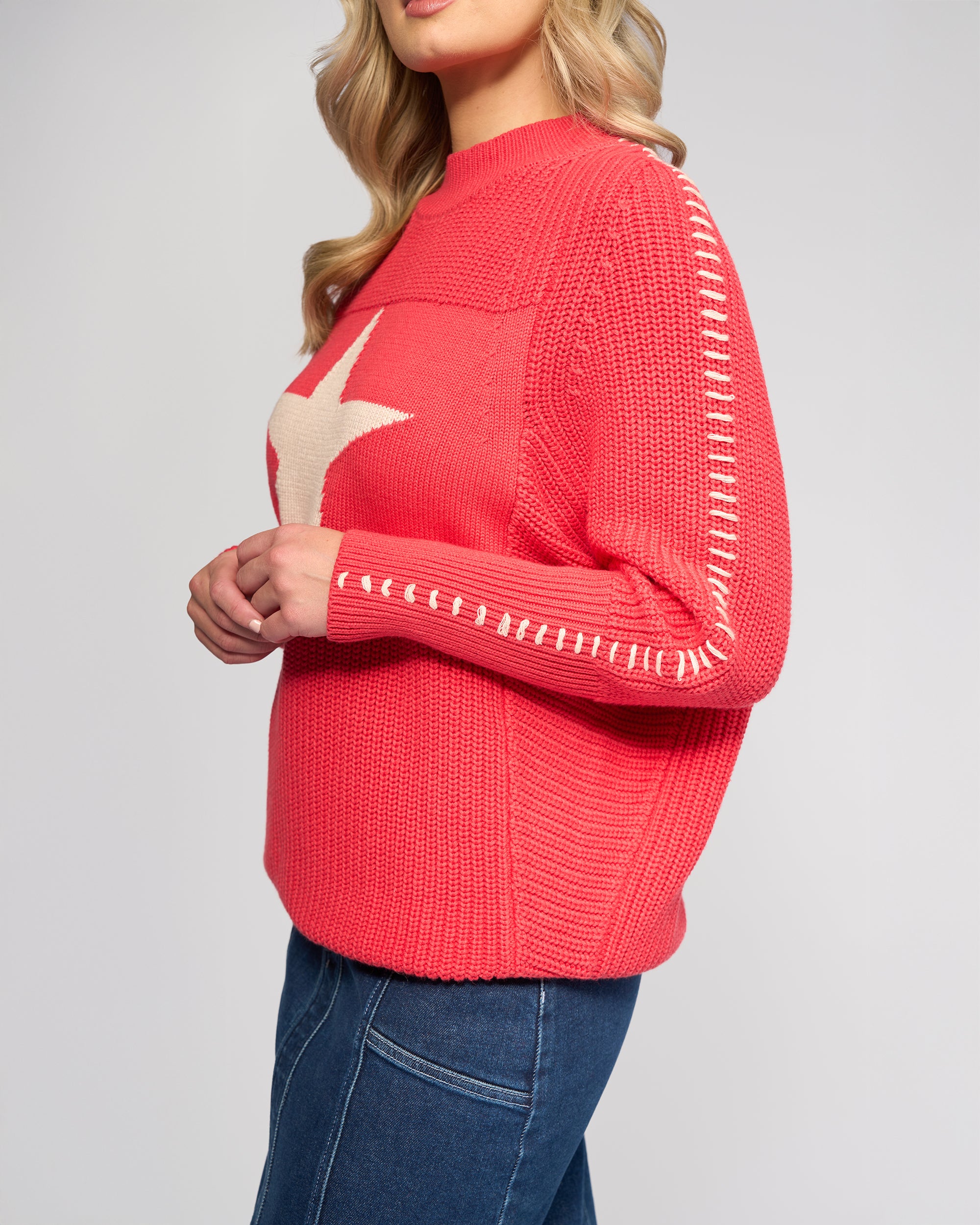 A woman with wavy blonde hair models the Vassalli Star Sweater - Coral, featuring white sleeve stitching and a bold front star, paired with blue jeans against a plain light background.