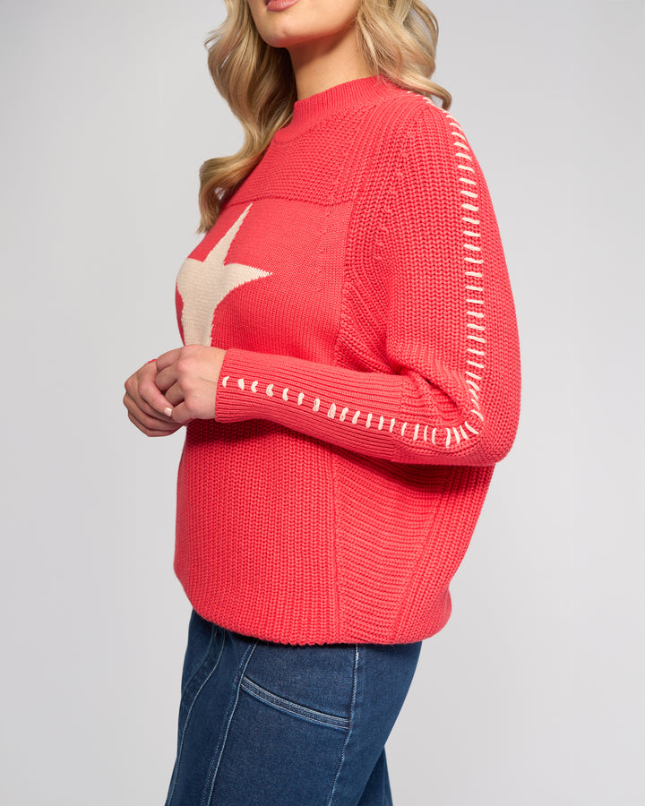 A woman with wavy blonde hair models the Vassalli Star Sweater - Coral, featuring white sleeve stitching and a bold front star, paired with blue jeans against a plain light background.